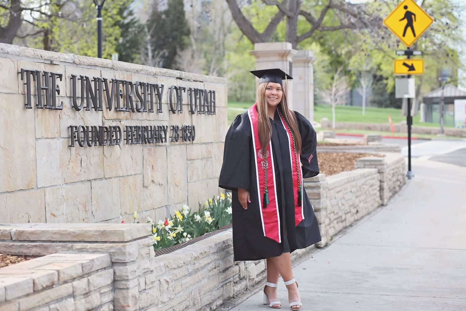 Graduate poses near University of Utah campus sign wearing graduation regalia.