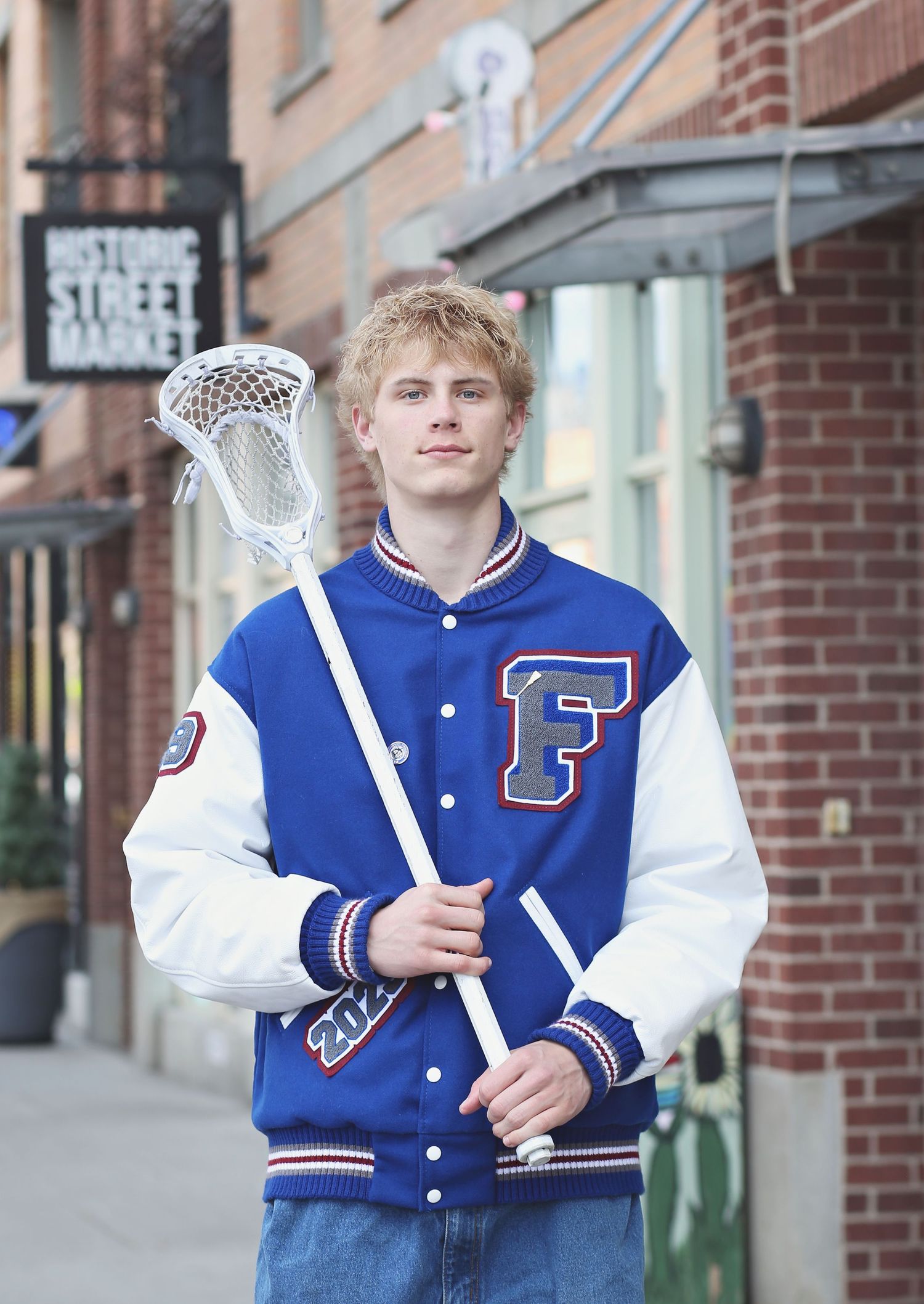 A student athlete in a blue and white varsity letterman jacket poses with a lacrosse stick against a brick building exterior.