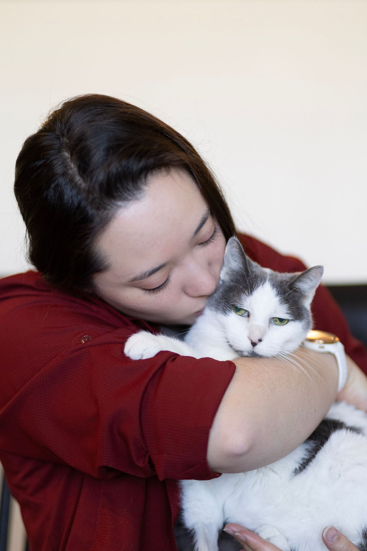 Woman in red hoodie kissing her gray and white cat while holding it in her arms indoors.