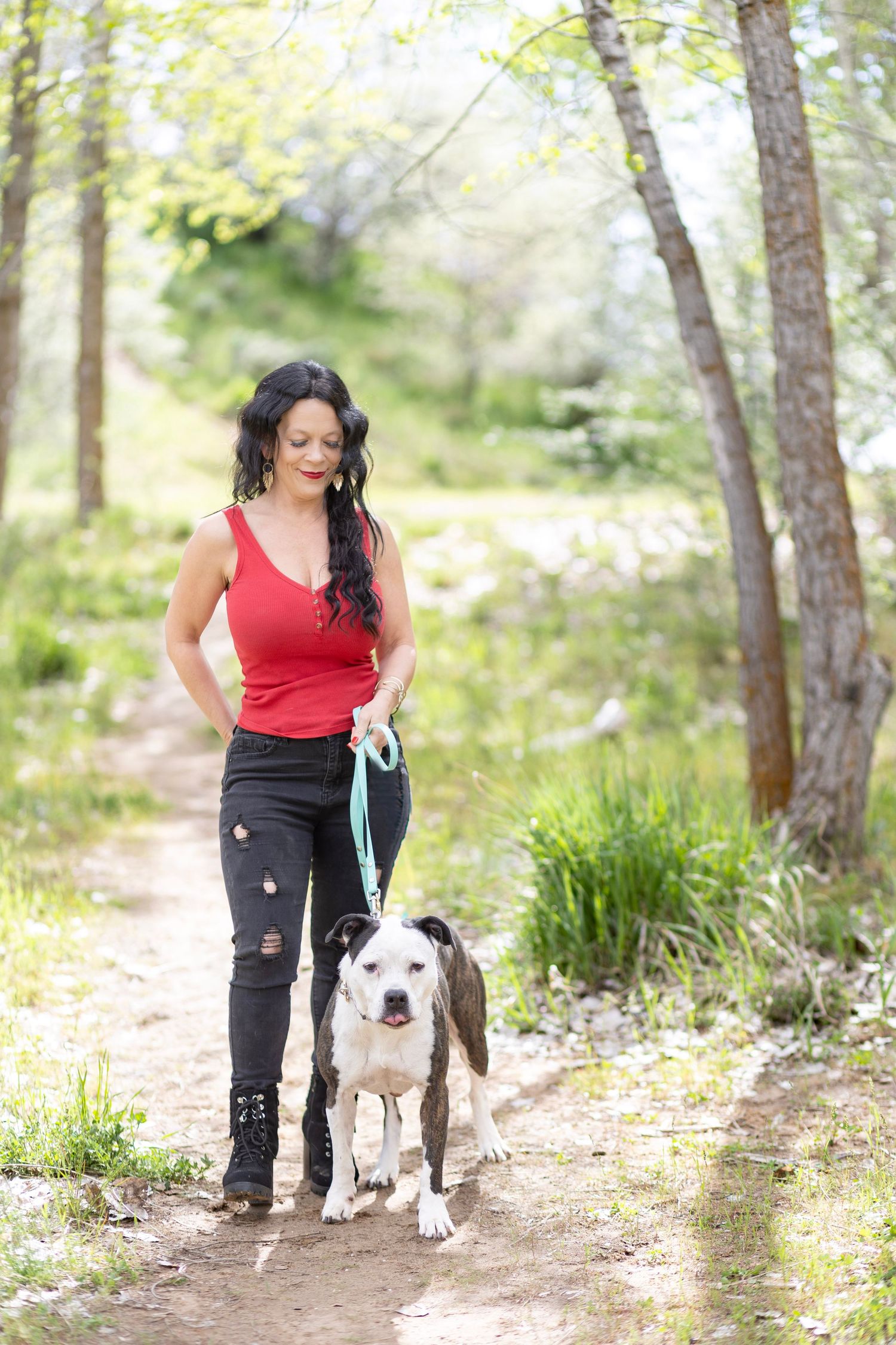 Woman in red tank top walking a black and white pit bull on a blue leash along a wooded trail.