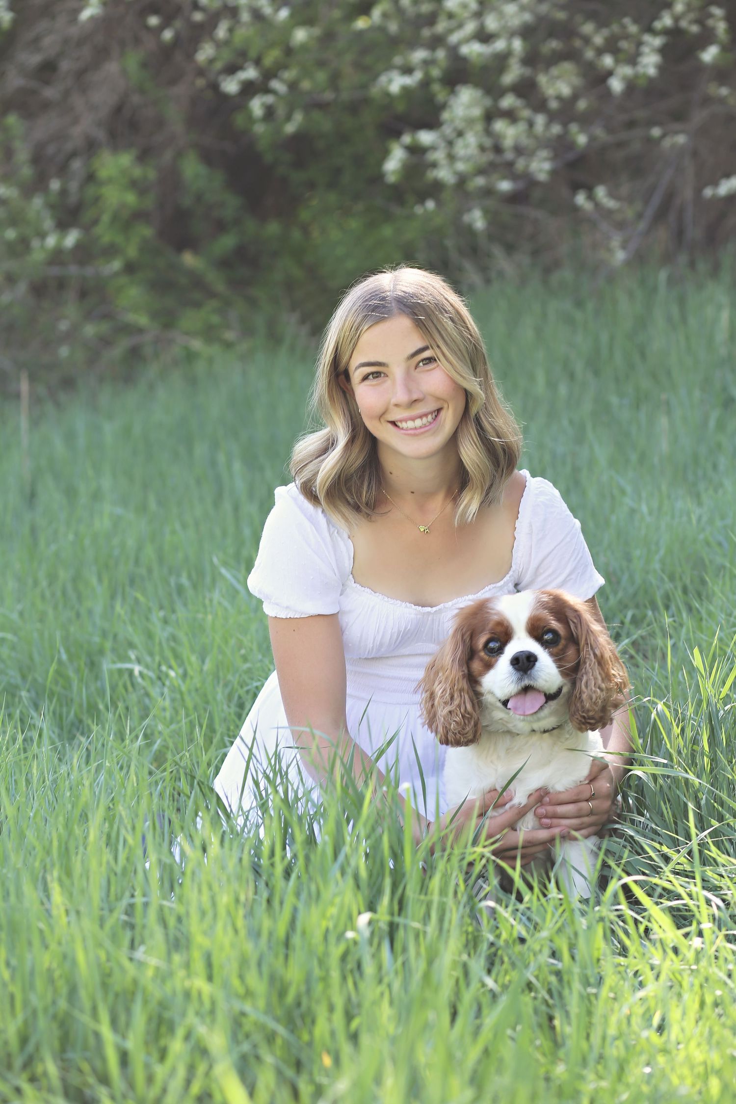 Woman in white dress sitting in tall grass with a small Cavalier King Charles Spaniel dog enjoying a sunny day.