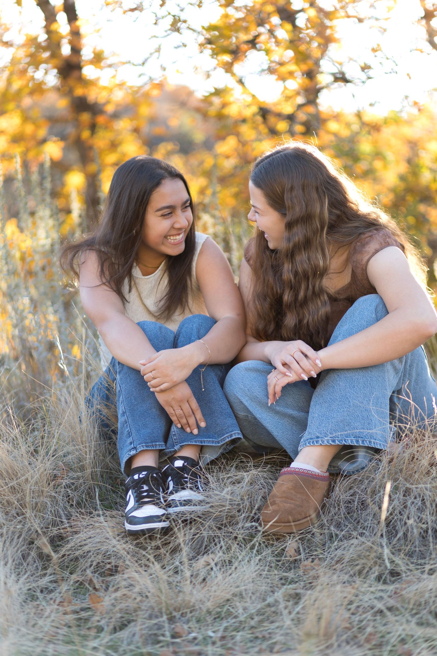 Two friends share laughter while sitting in tall grass during golden hour sunset.