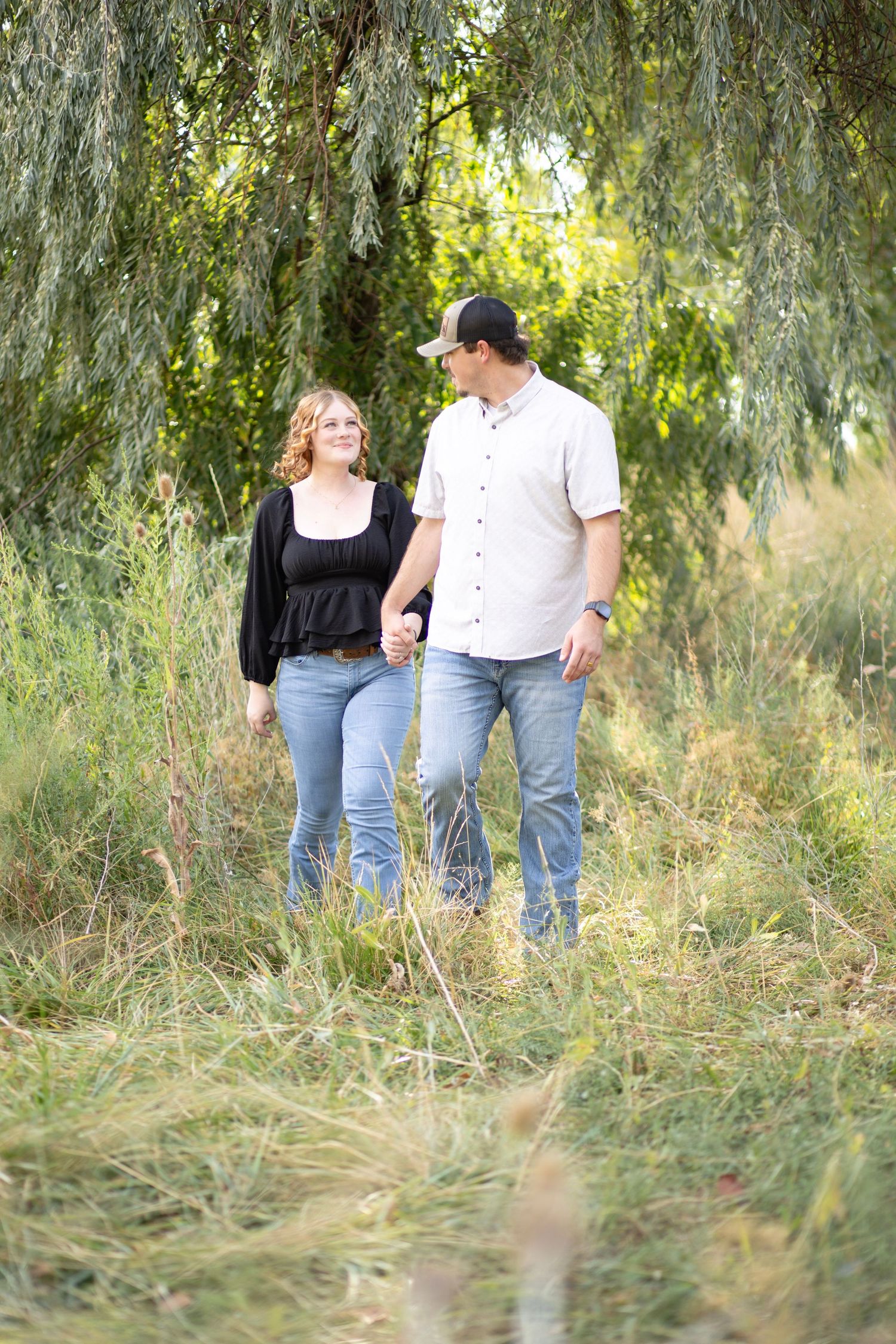 Couples walk through a scenic area with willow trees and tall grass during a photo session.