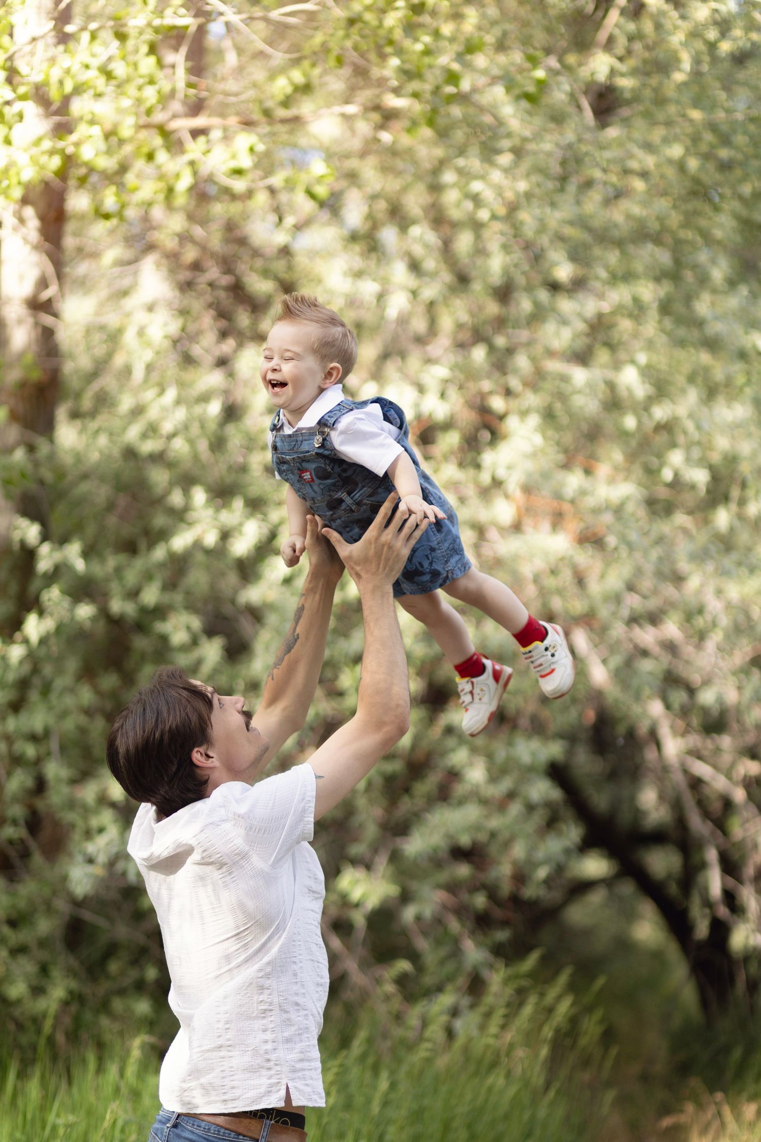 A joyful father lifts his laughing toddler high in the air outdoors, surrounded by lush green trees.