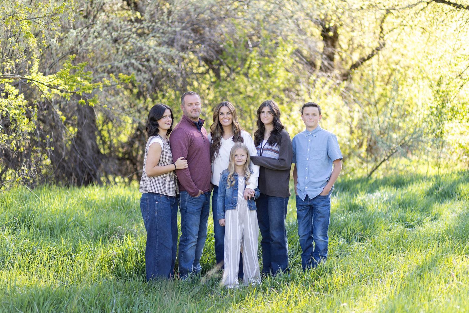 Family of six posing outdoors in green meadow with blooming trees, casual attire, portrait photography.