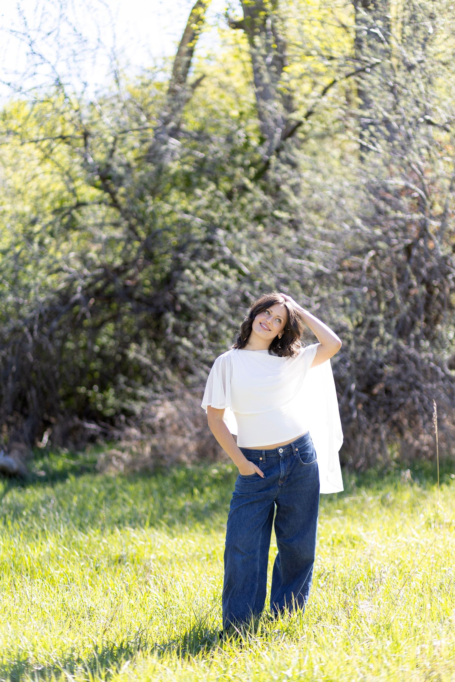 Young woman in white tee and wide-leg jeans poses outdoors in sunny green field with trees.