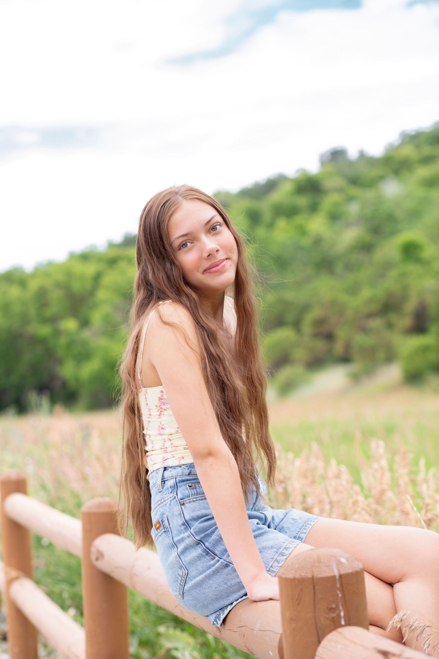 Casual outdoor portrait sitting on wooden fence with denim shorts and white top in a meadow setting.