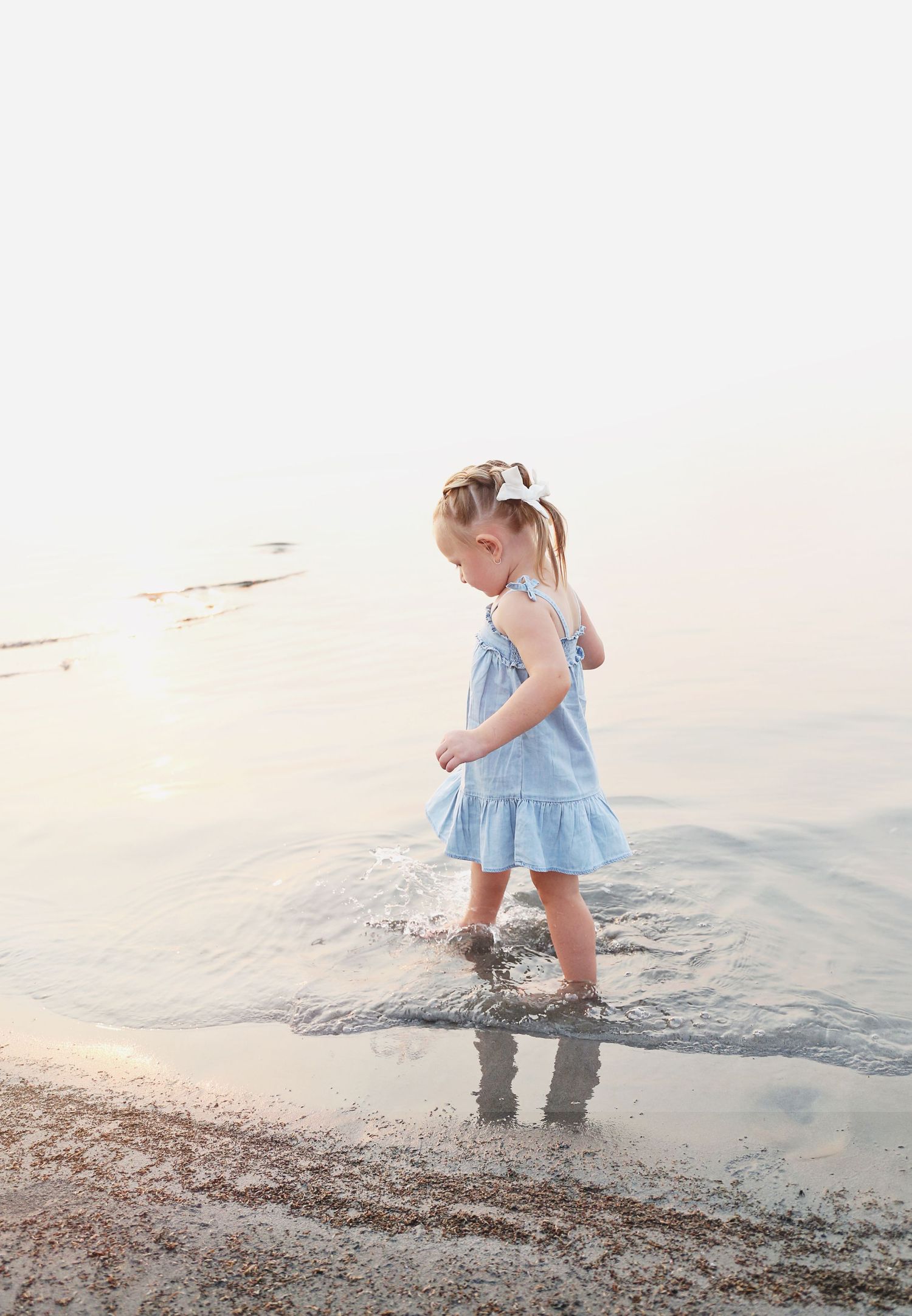 A child in a blue dress plays at the water's edge during sunset.