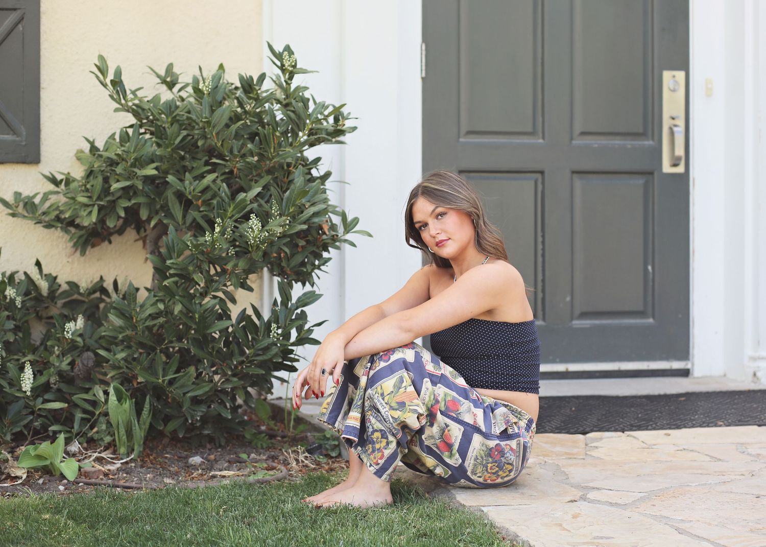 A person sits on a doorstep in a floral dress and beige flats while holding a glass of wine.