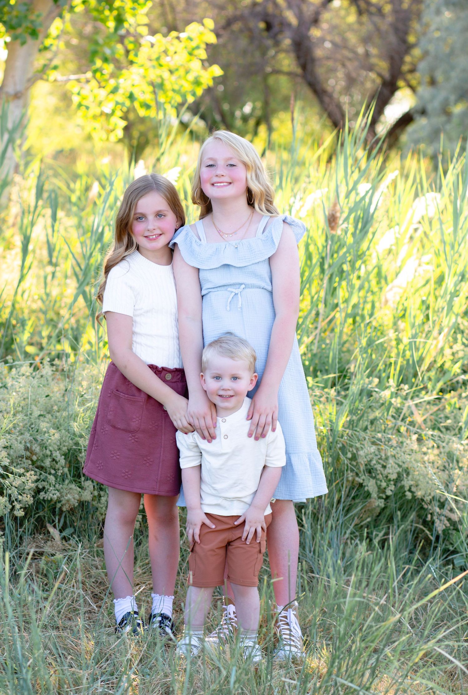 Three siblings pose together outdoors against a backdrop of tall grass and sunlit trees.