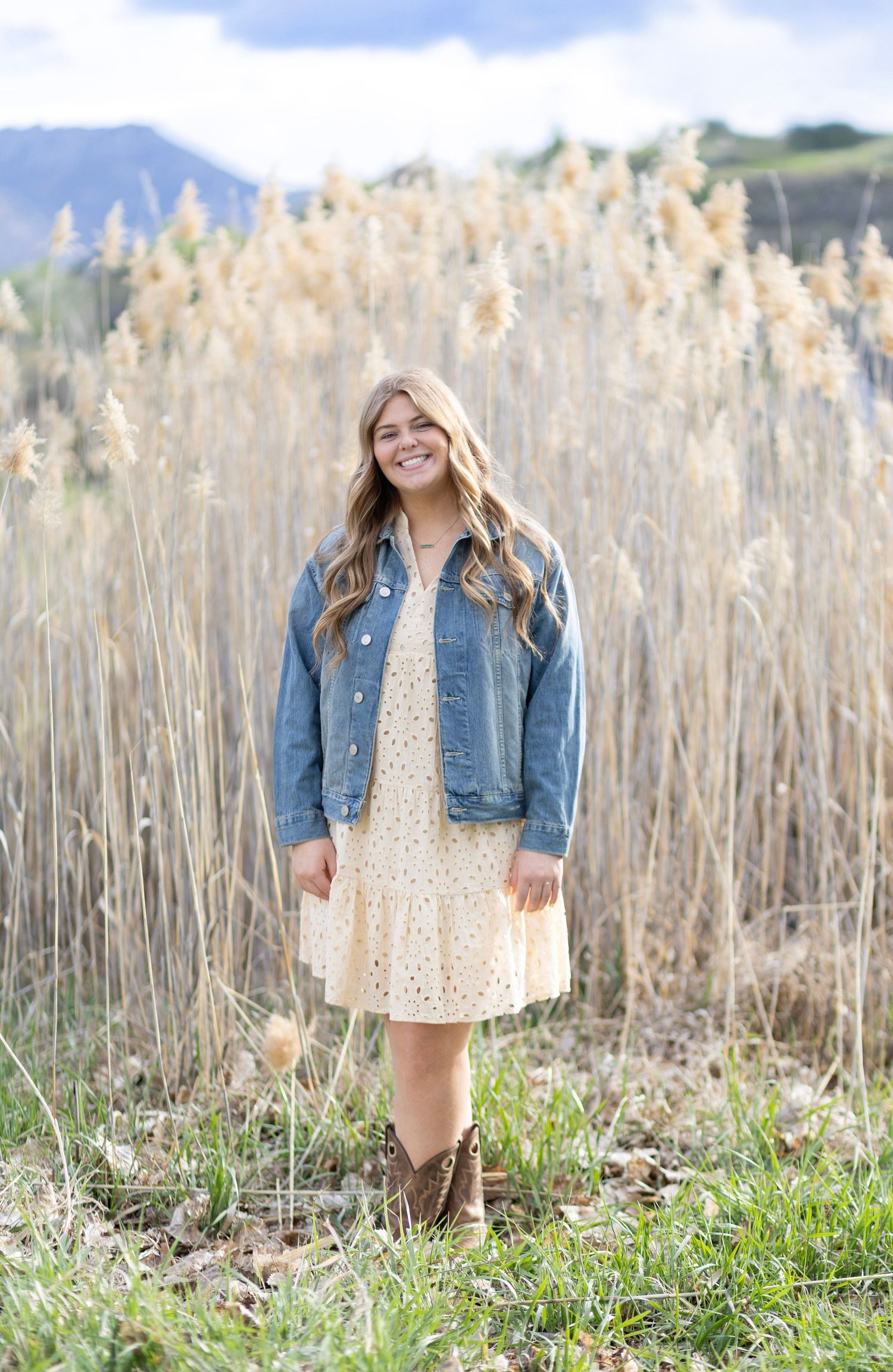 Smiling young woman in denim jacket and floral dress standing in a field of tall golden grasses with mountains behind.