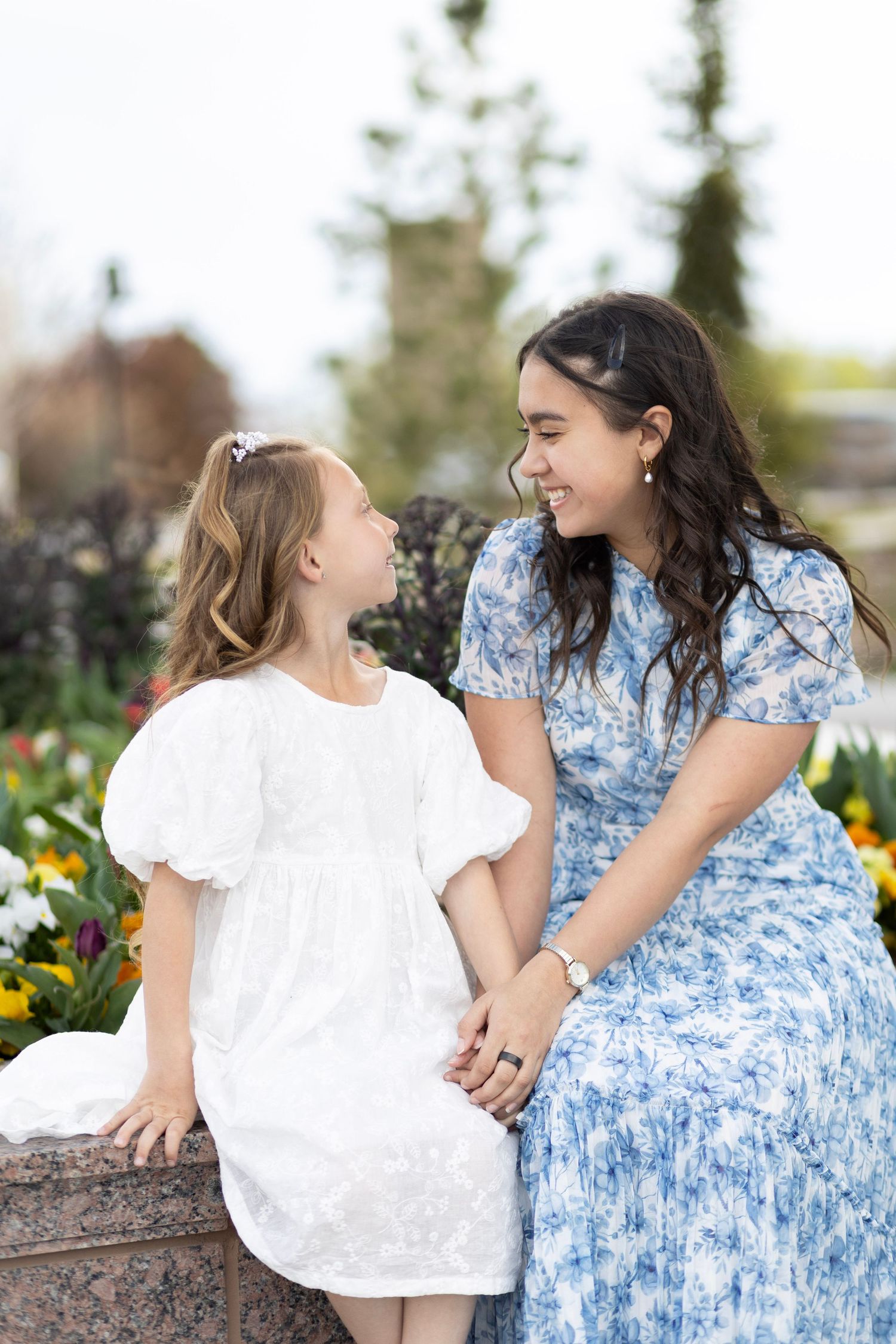 A smiling teenage girl in a blue floral dress sits with a young girl in white dress among colorful flowers in a garden.