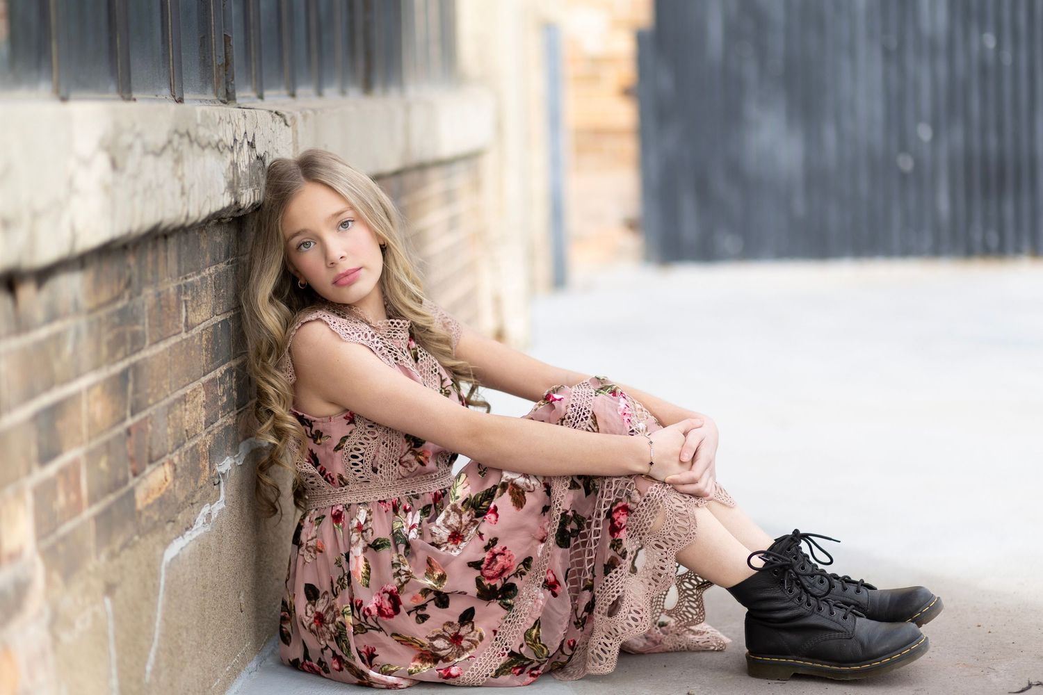 Young girl in floral dress and black boots sitting against a brick wall outdoors.