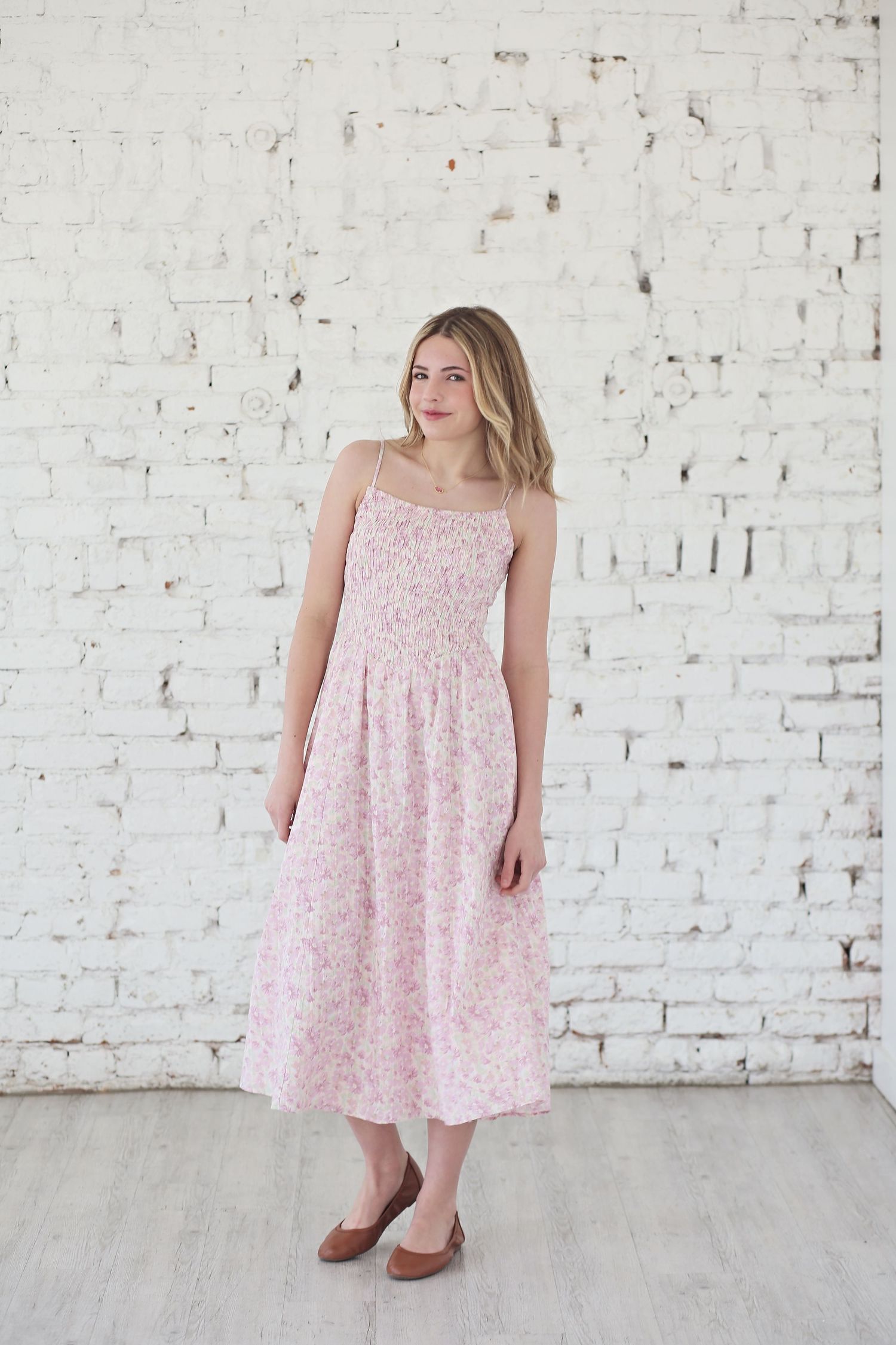 A person in a pink floral sundress poses against a white brick wall.