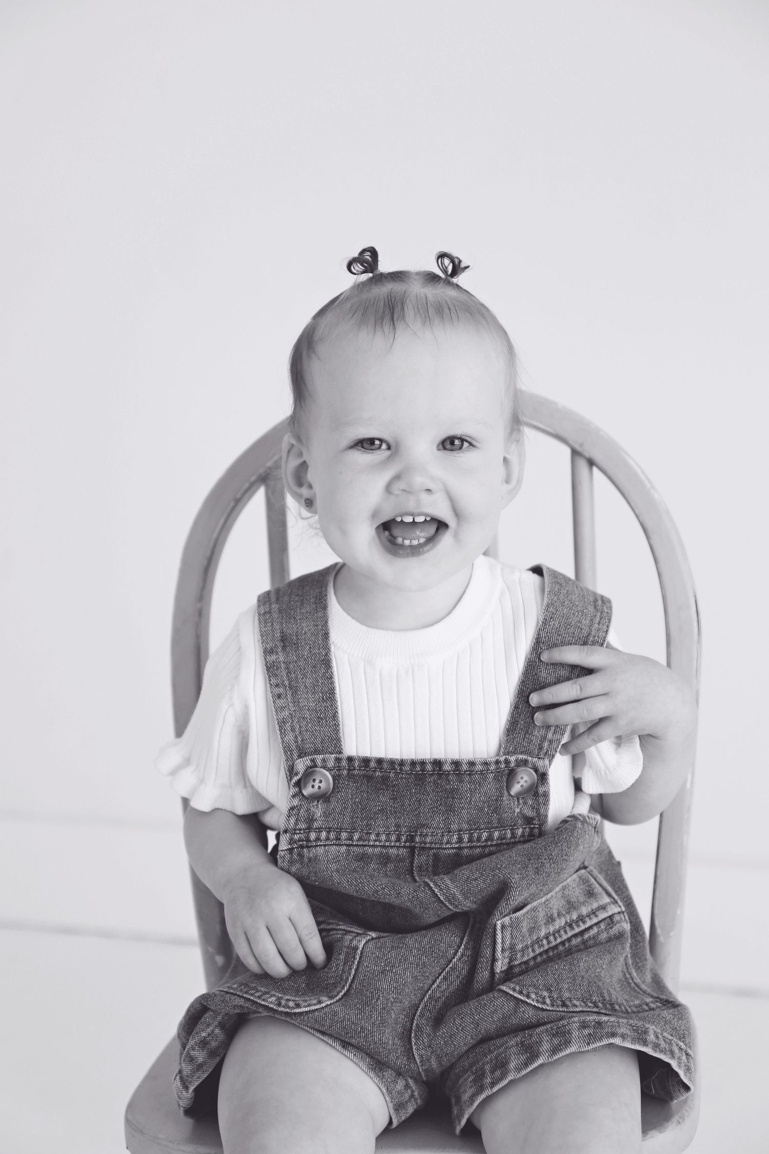 Toddler in denim overalls sits smiling in a light blue wooden chair.