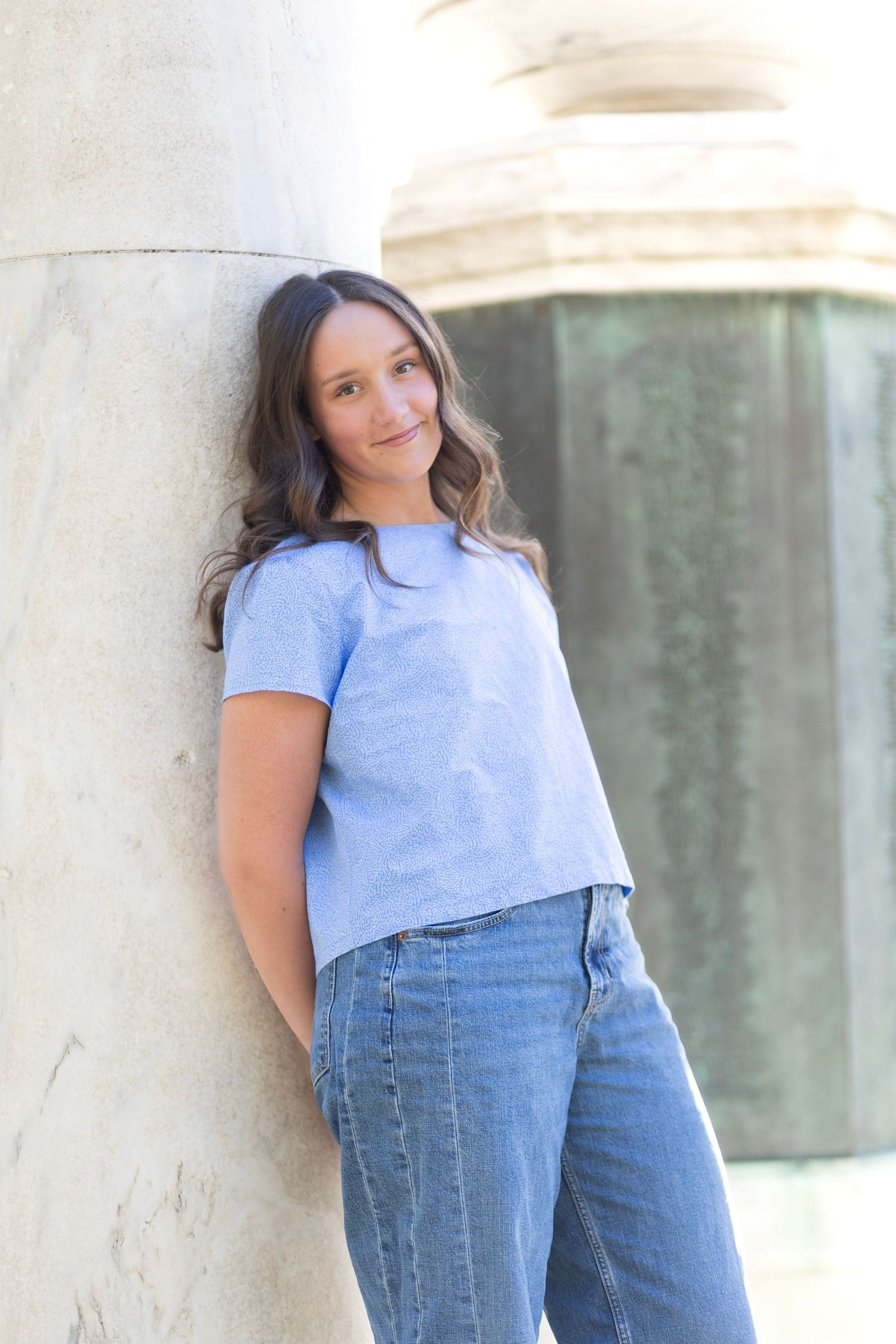 Young woman in blue t-shirt and jeans leaning against a stone column outdoors, smiling softly.