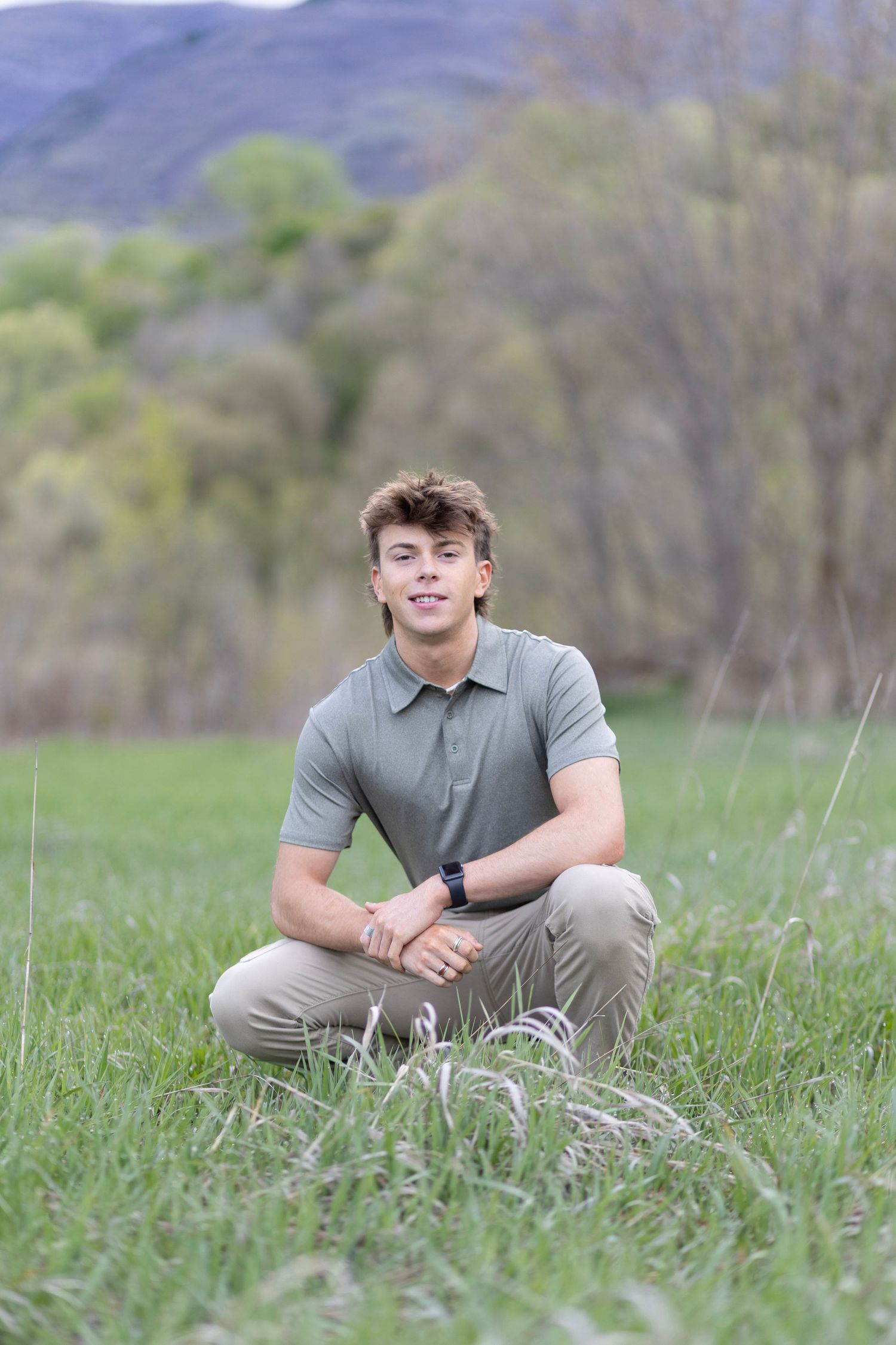 Young man in olive polo shirt sitting cross-legged in green meadow with mountain backdrop.