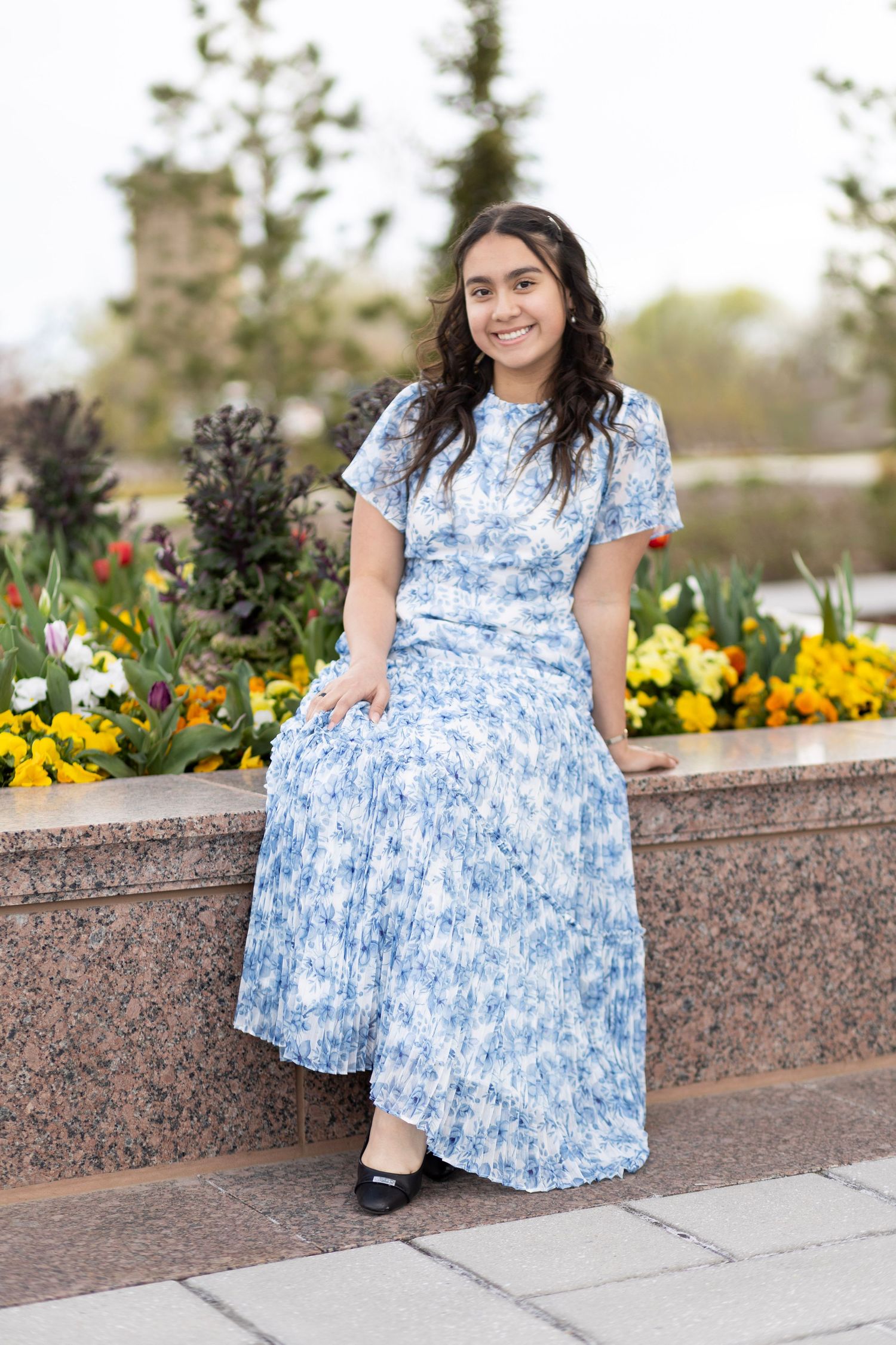 Young woman in blue floral midi dress poses outdoors near flower garden, smiling gracefully.