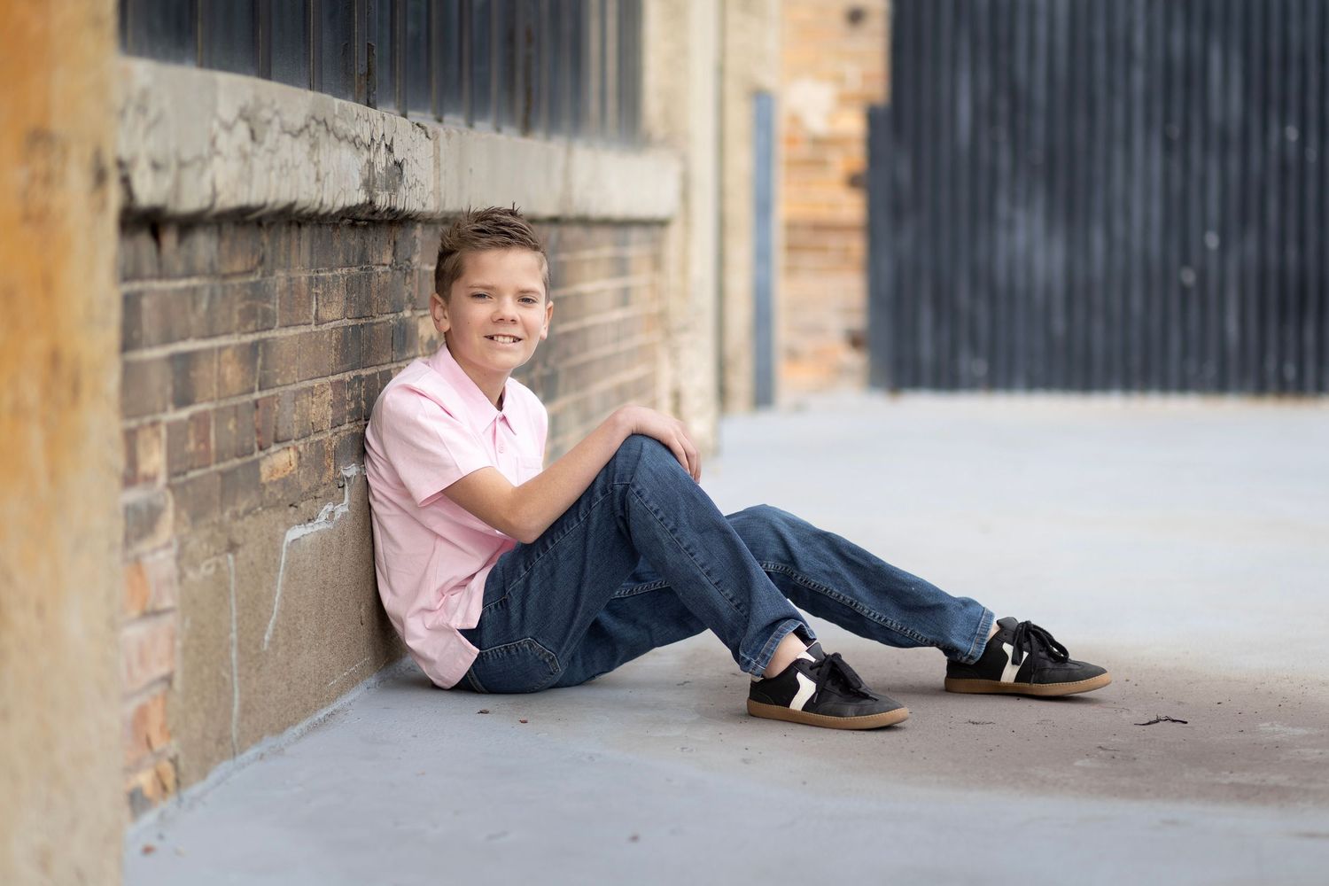 Smiling boy in pink shirt and jeans sitting against a brick wall outdoors.