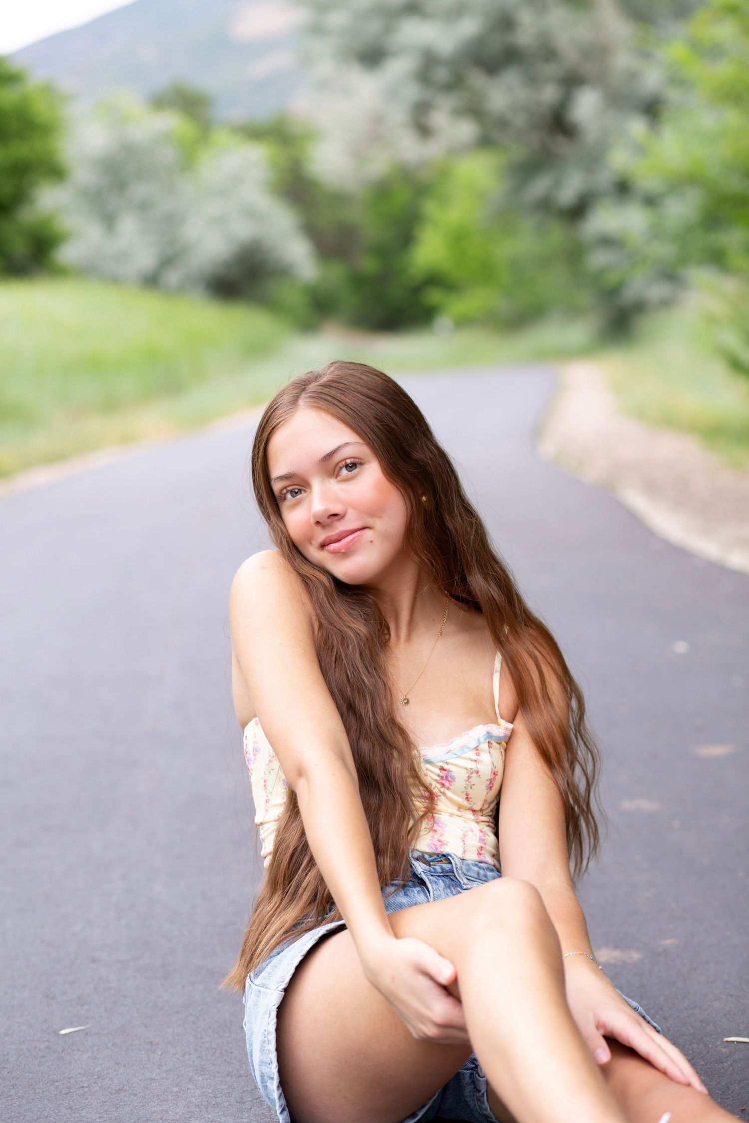 A summer portrait on a mountain road featuring long brown hair, denim shorts and a floral top against a blurred nature backdrop.