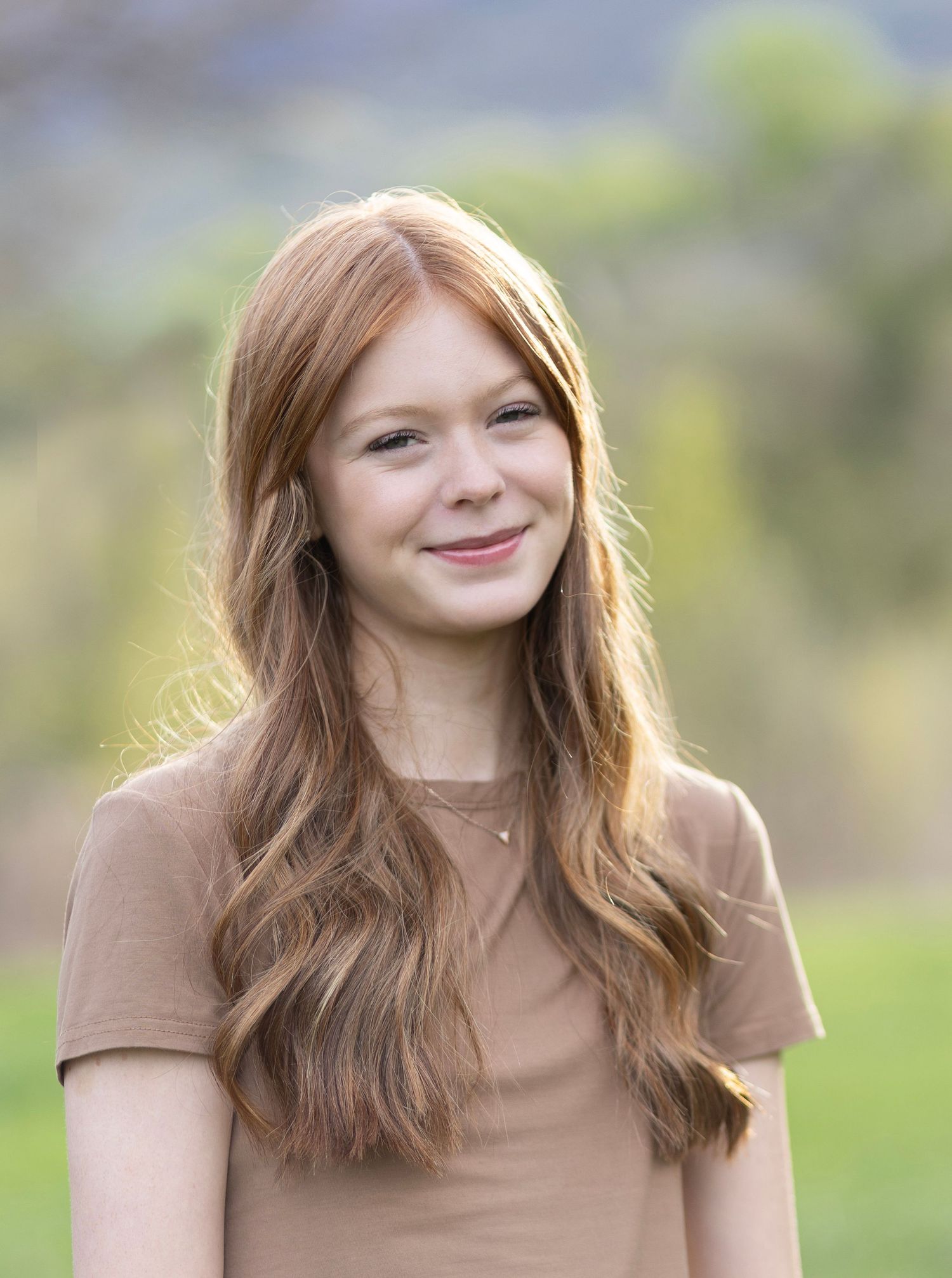 Smiling teenage girl with long auburn hair wearing a tan t-shirt outdoors with green hills in background.