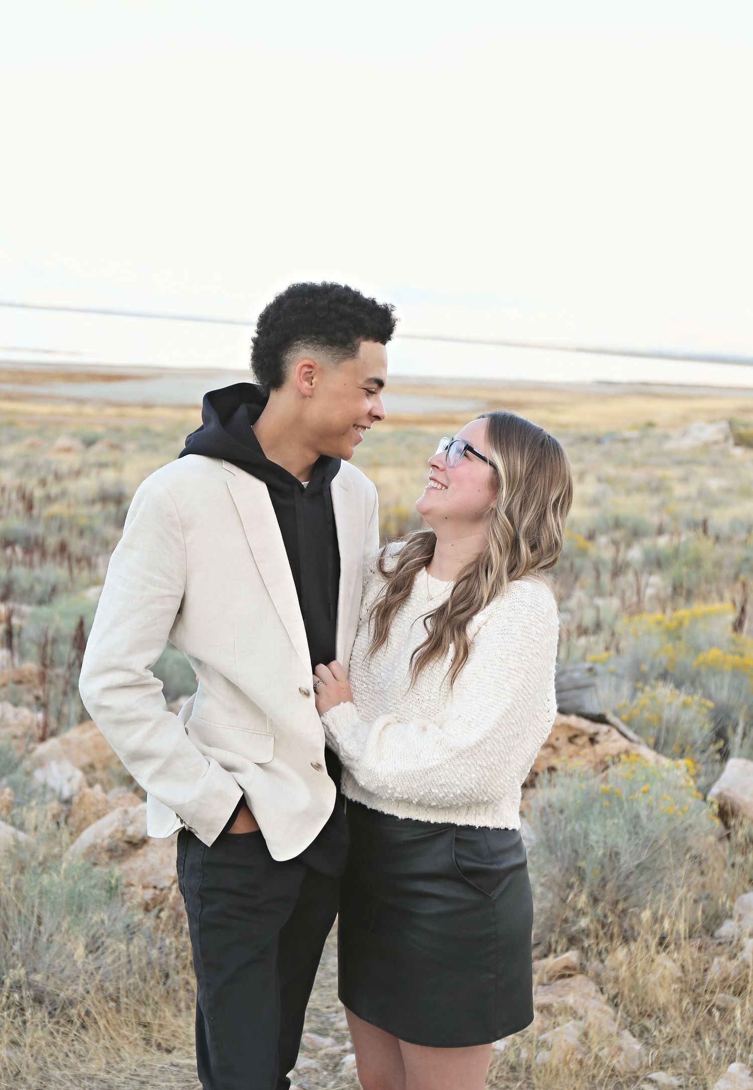 A couple laughs together in a desert setting with brush and mountains in background.