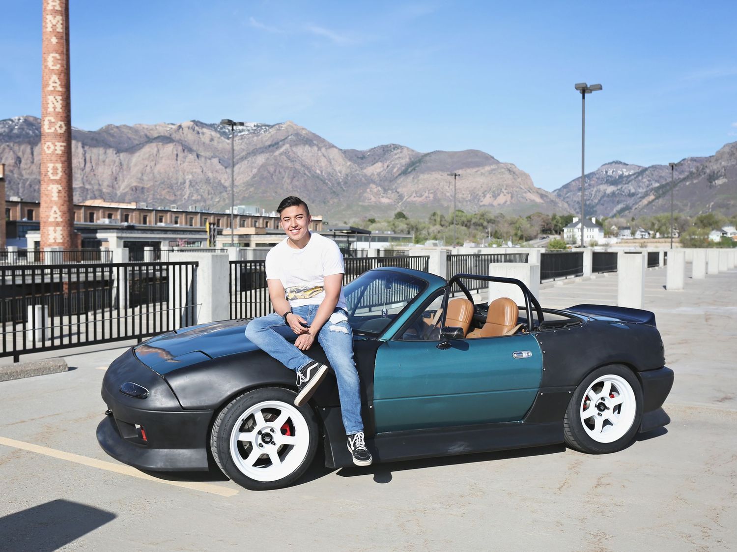 Person poses with dark blue sports car in front of mountain landscape.