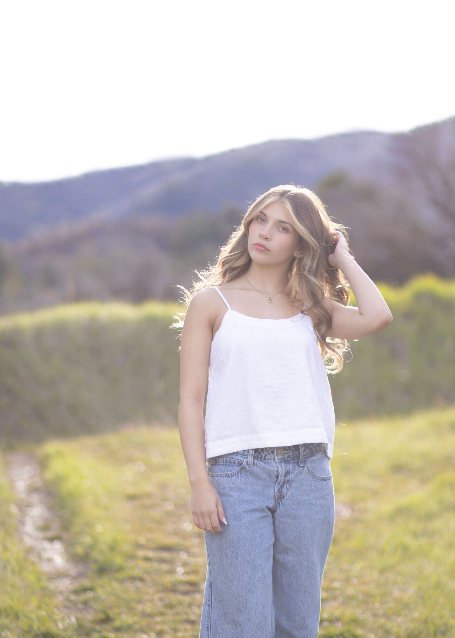 Young woman in white camisole and jeans poses in a sunlit green field with mountain backdrop.