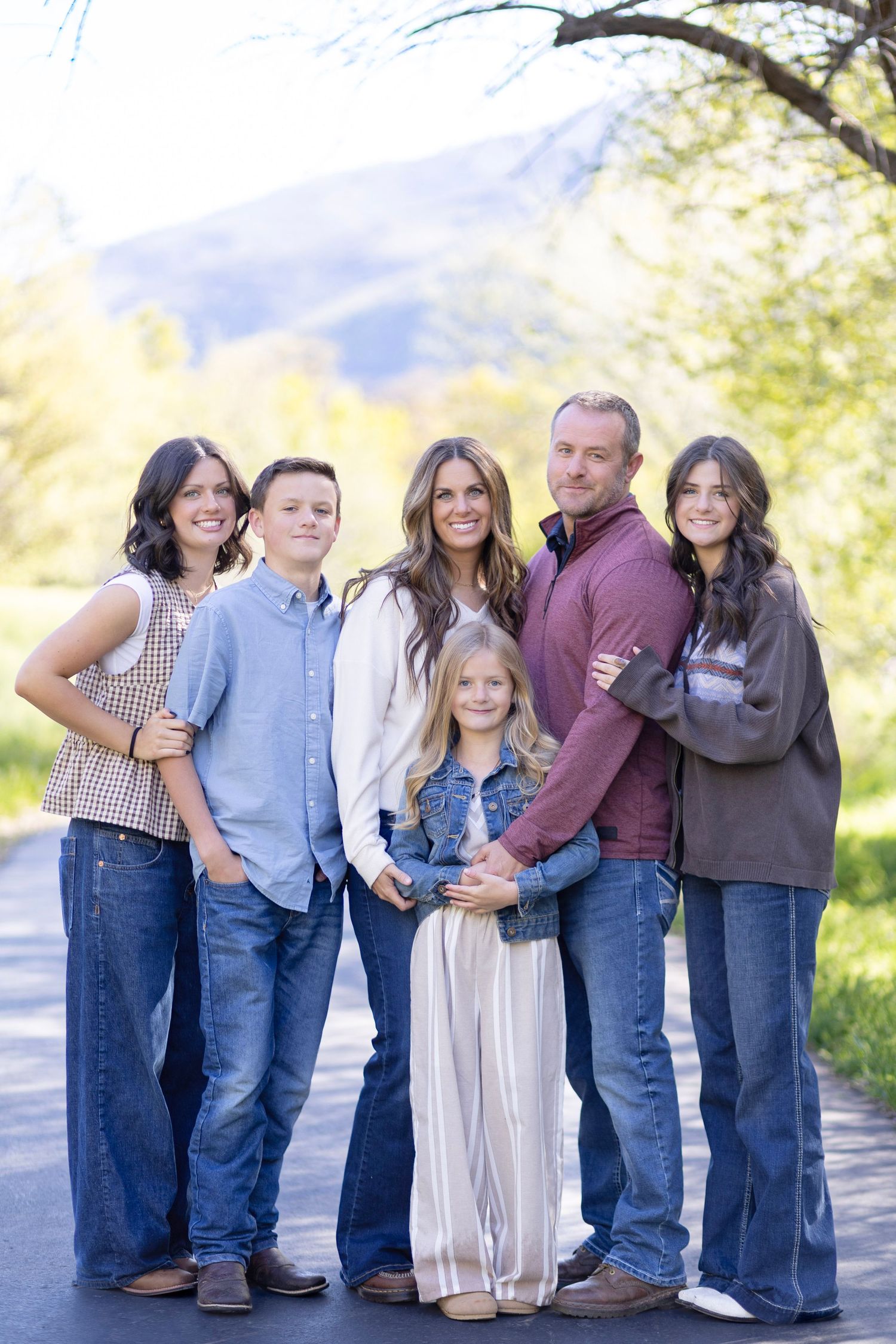 Family of six posing together on a scenic tree-lined road with mountain views in casual denim outfits.