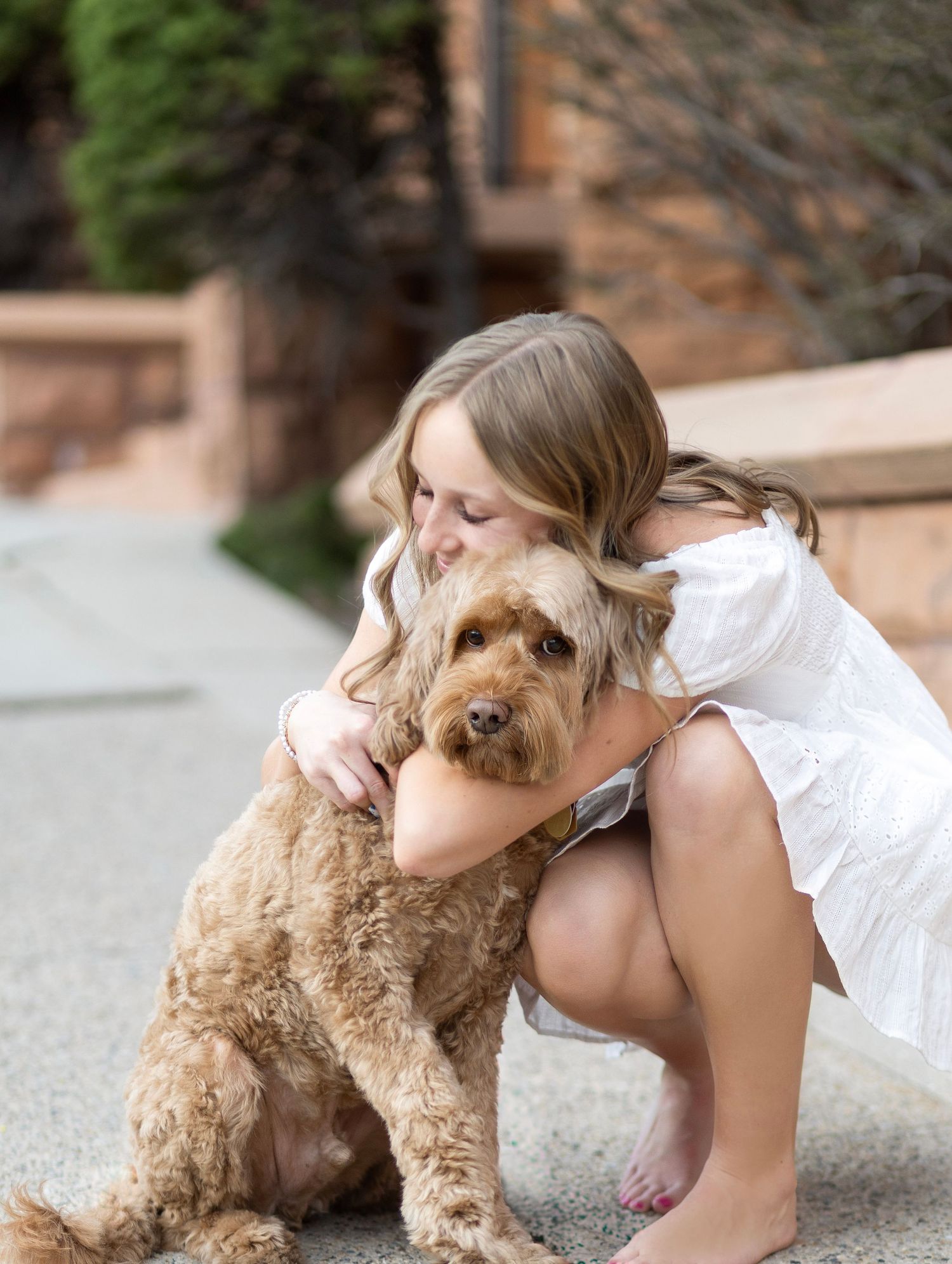 Young girl in white dress hugging a fluffy golden doodle dog outdoors on a sunny day.