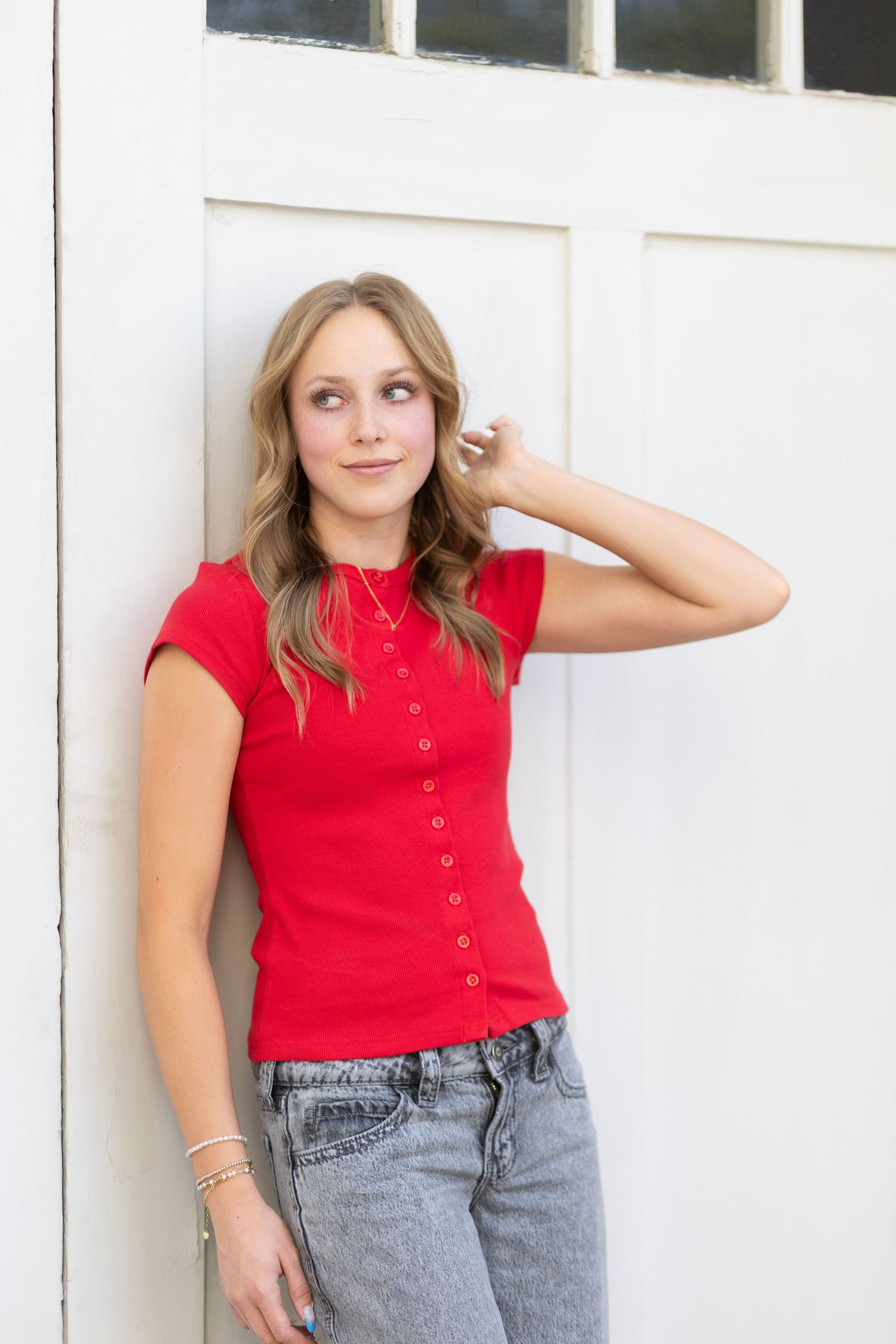 Young woman in red button-up top and jeans leaning against white door, smiling thoughtfully.