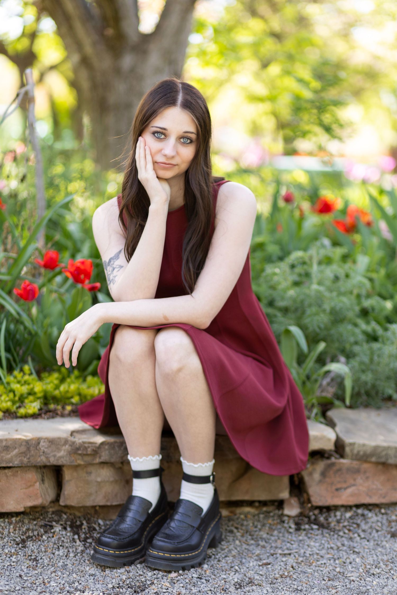 Young woman in burgundy dress and black loafers sitting pensively on stone border among colorful tulips.