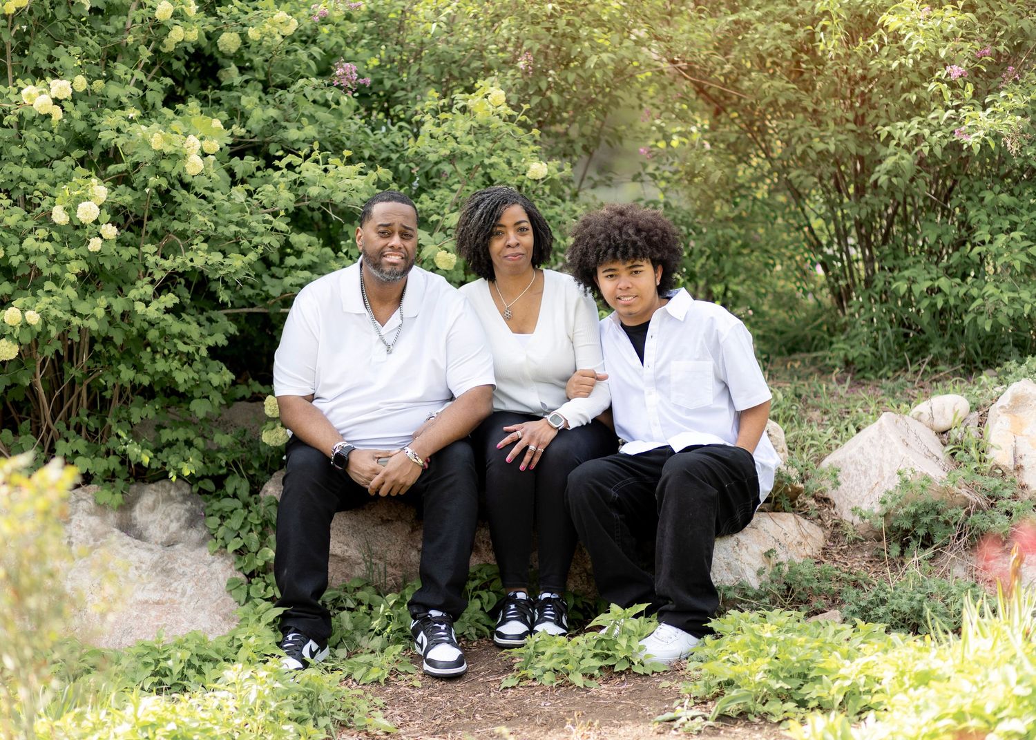 Family of three sitting on rocks in a lush garden, wearing white and black outfits, smiling outdoors.