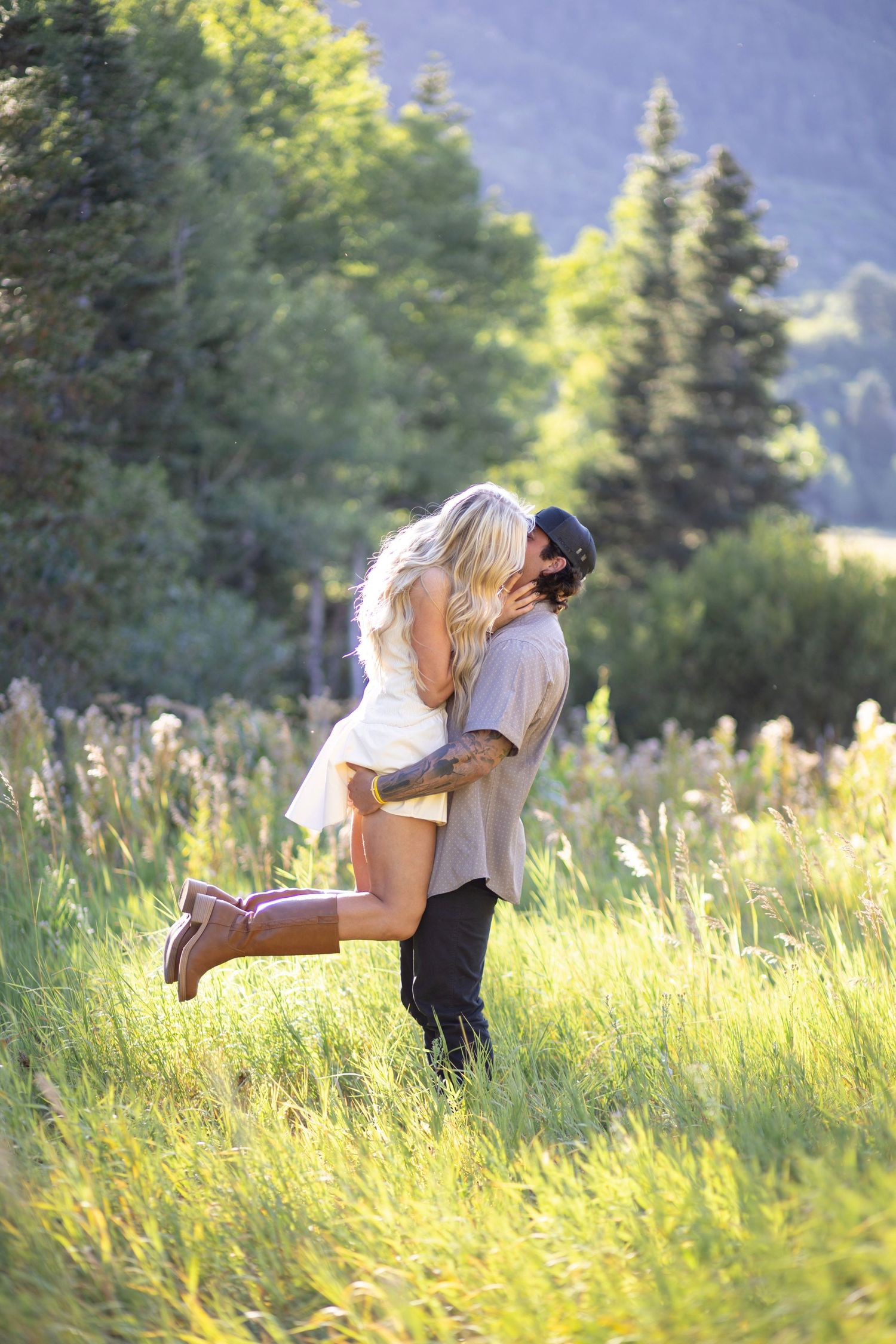Playful couple embracing in a golden meadow with evergreen trees and mountains behind them.