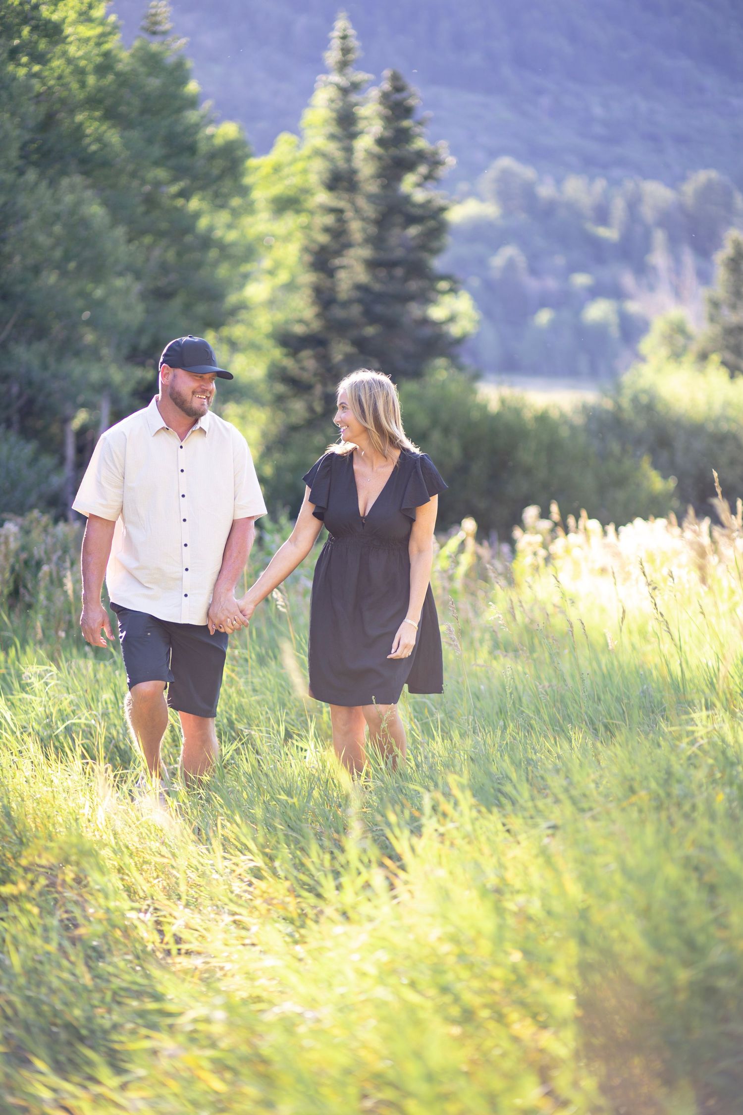 Couple walking through sunny meadow with mountains in background.