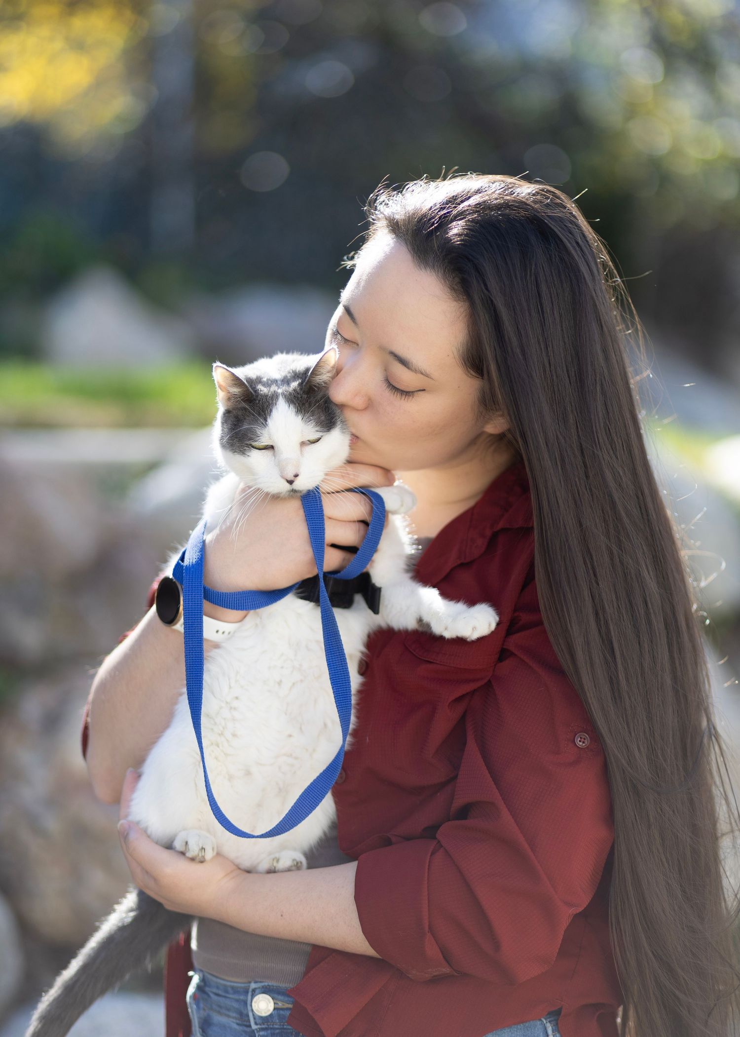 Young woman with long hair kissing a gray and white cat on a blue leash outdoors.