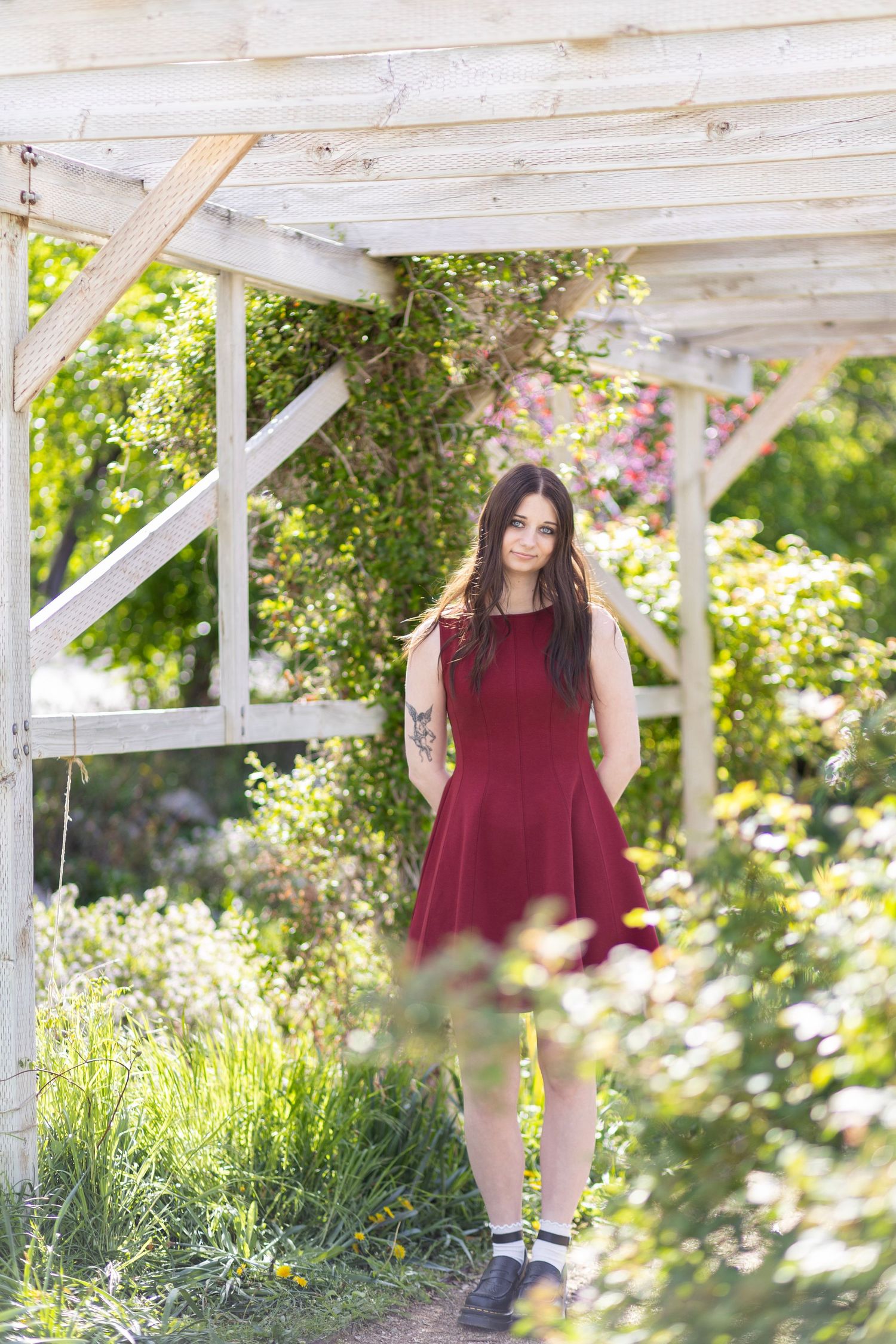 Young woman in burgundy dress standing under white wooden pergola surrounded by lush green garden foliage.