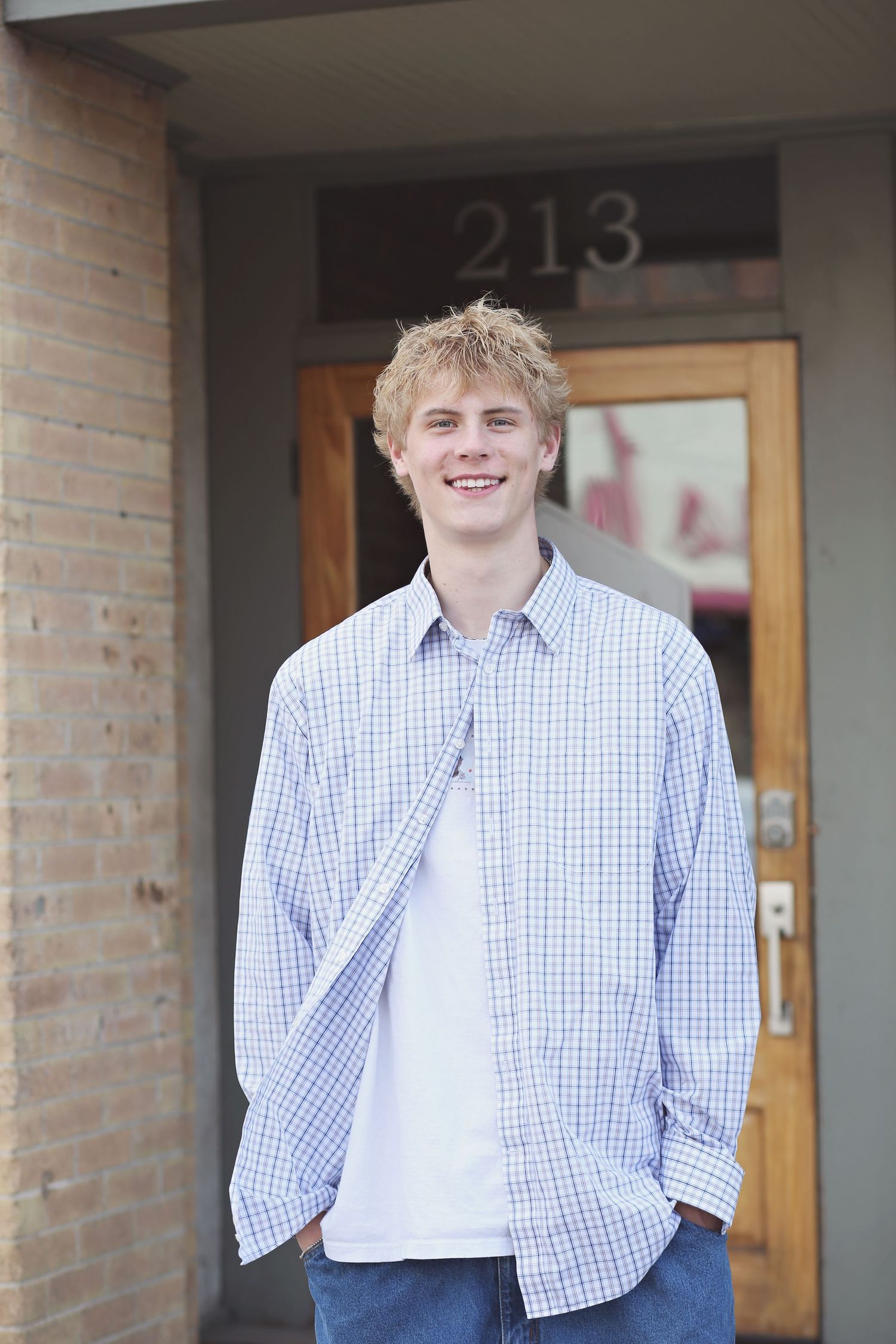 A casual portrait in front of room 213 wearing a checkered button-down shirt.
