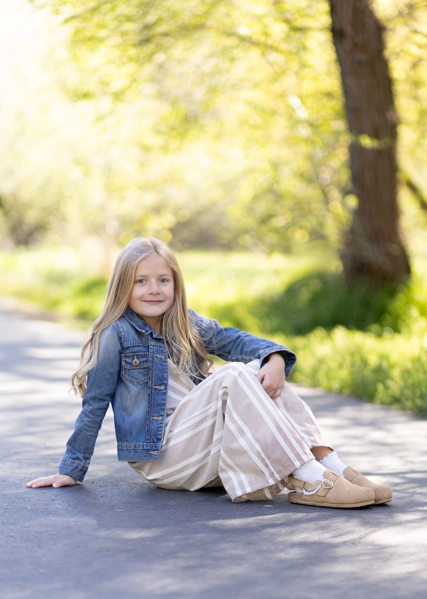 Young girl in denim jacket and striped skirt sitting on a sunlit path surrounded by lush green trees.