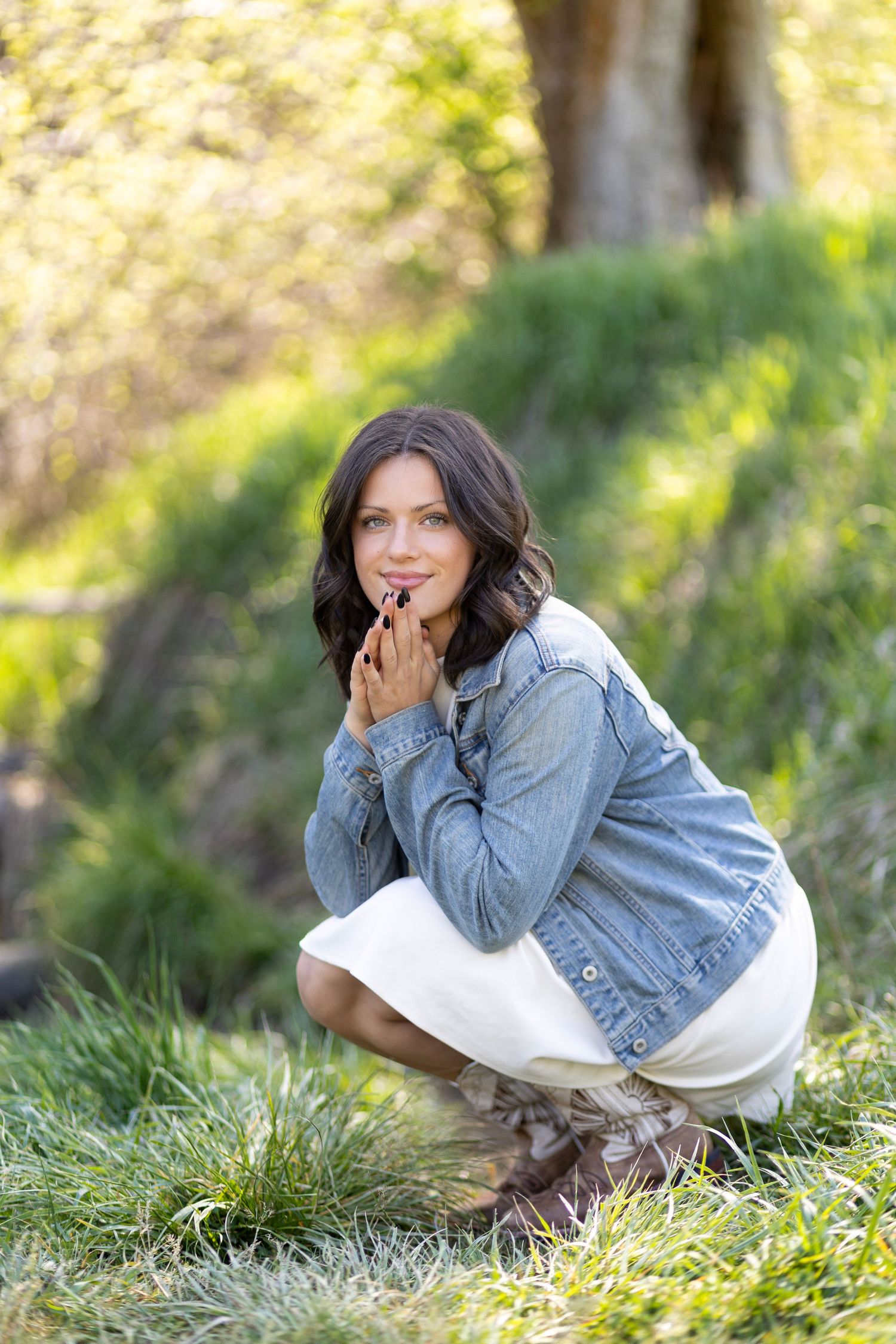 Woman in denim jacket and white skirt crouching outdoors in lush green park setting, smiling with hands clasped.