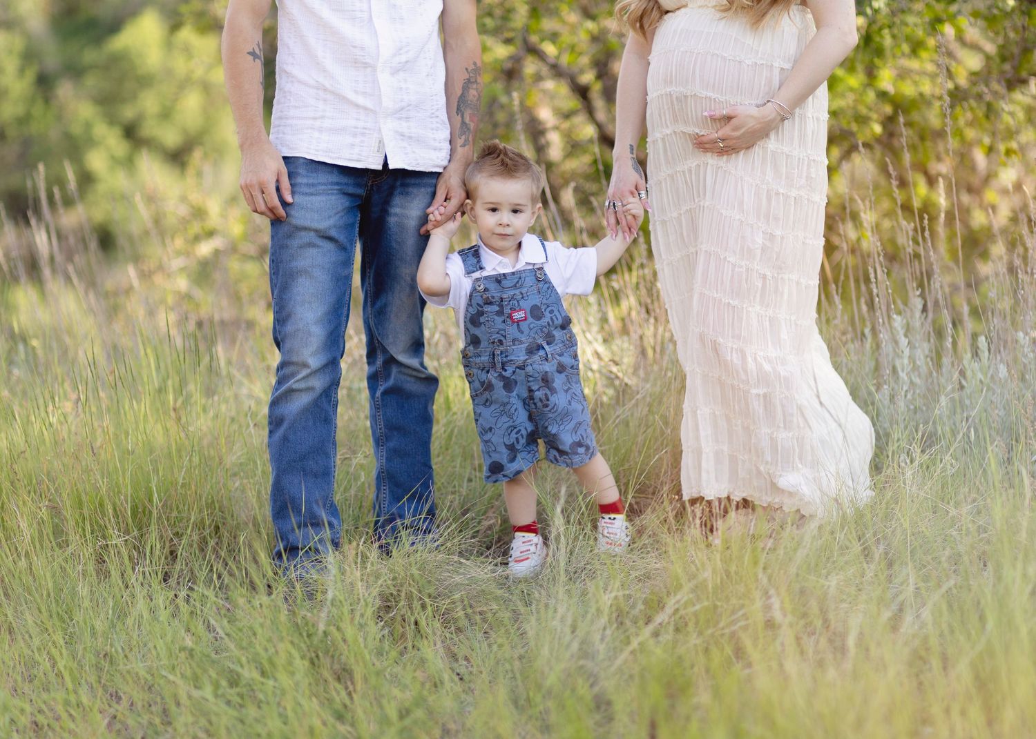 Pregnant woman and man holding toddler boy's hands in a sunny green meadow during a maternity photo session.