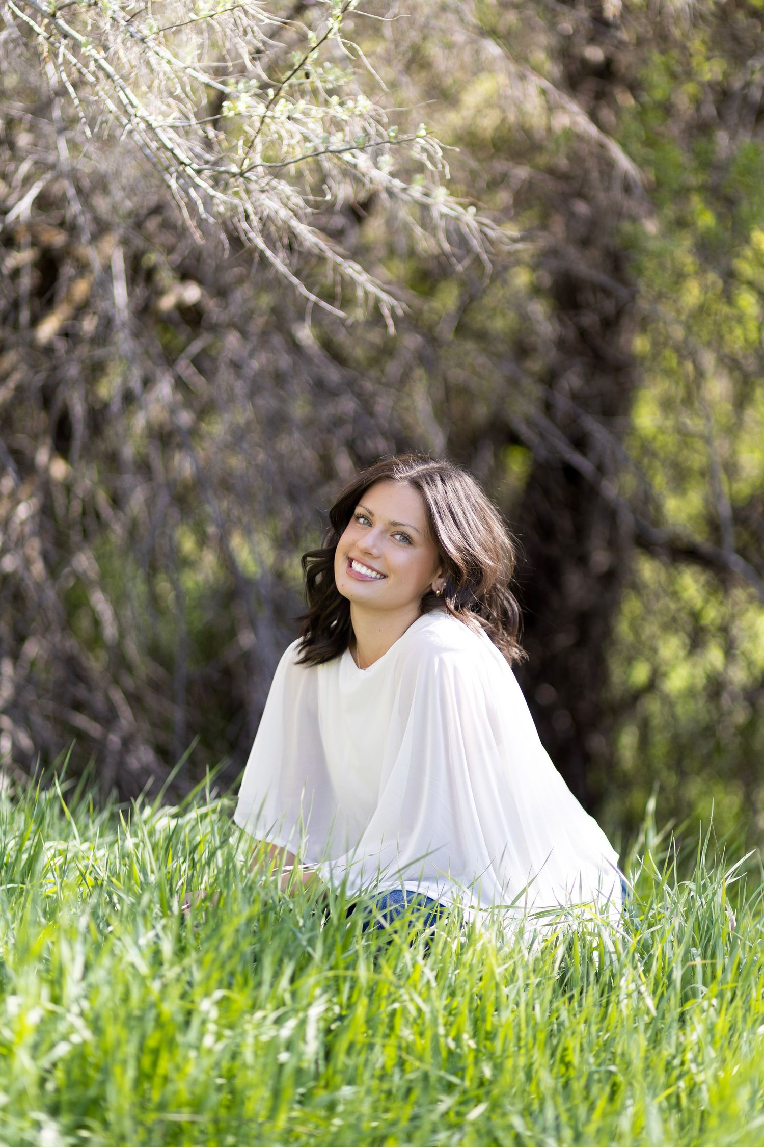 Smiling brunette woman in white top sitting in lush green grass outdoors near trees on a sunny day.
