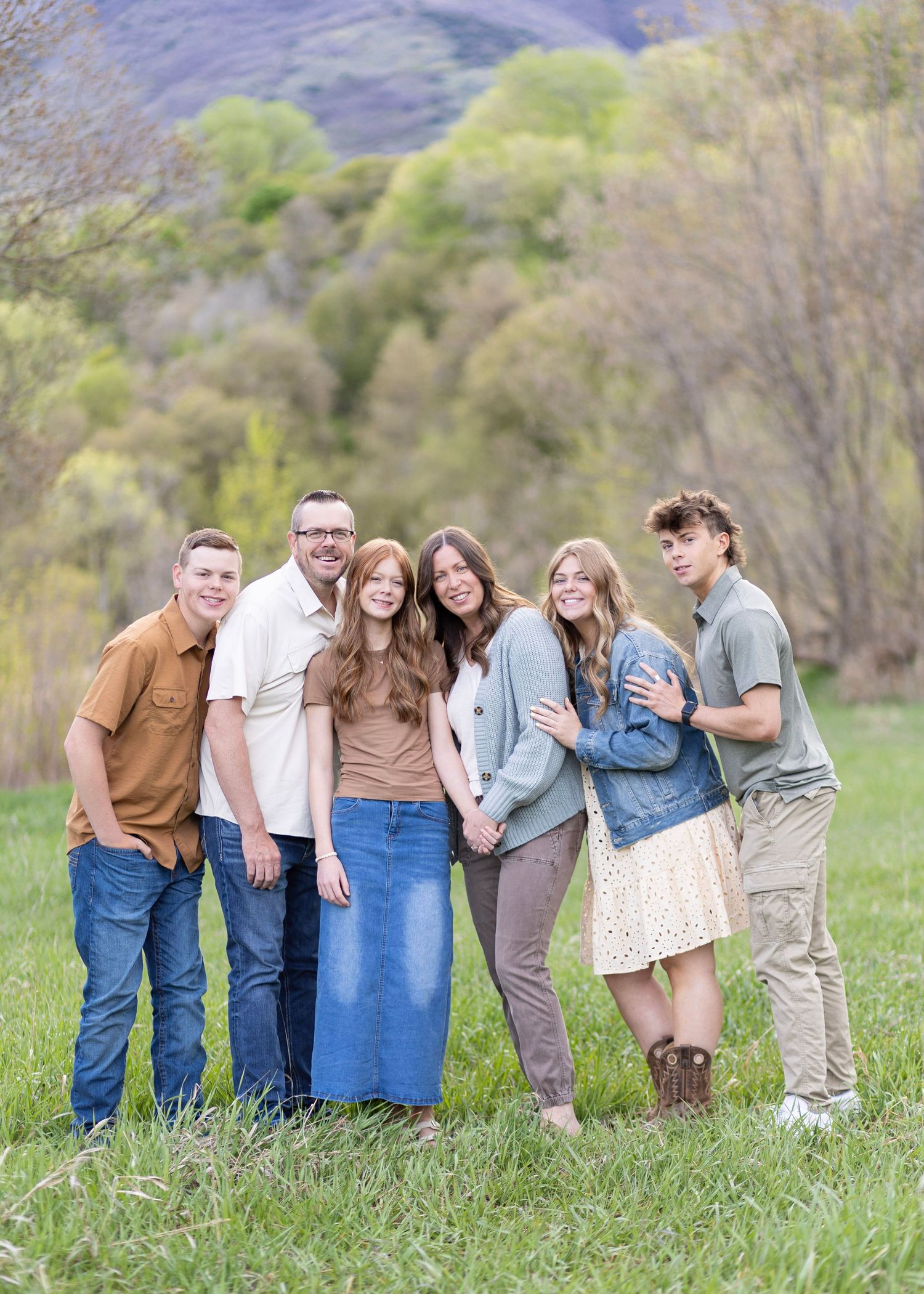 Family of six posing together in a green meadow with tree-covered hills in the background on a spring day.