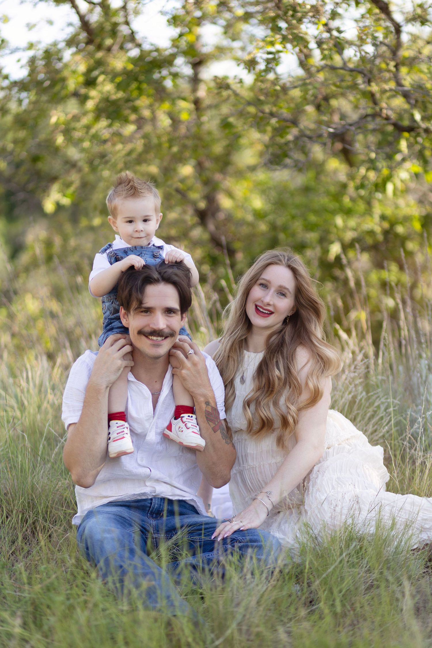 Happy young family with toddler on dad's shoulders sitting in green meadow, smiling outdoors.