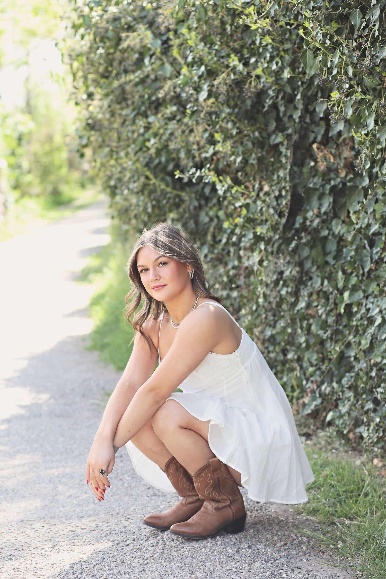 A person in a white dress and brown boots sits casually on a gravel path near green hedges.