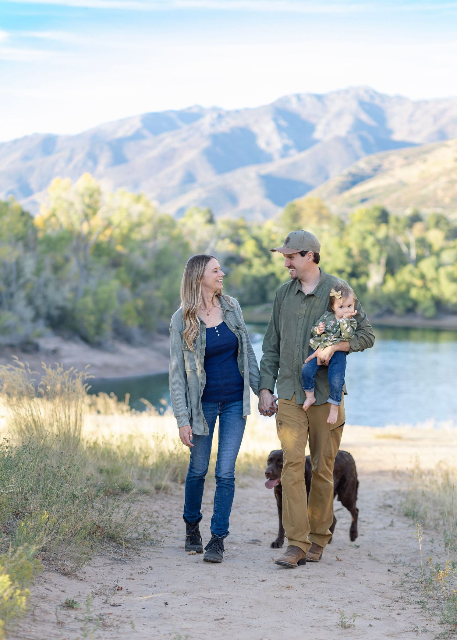 Family walking along lakeside path with mountains in background during sunset photo session.