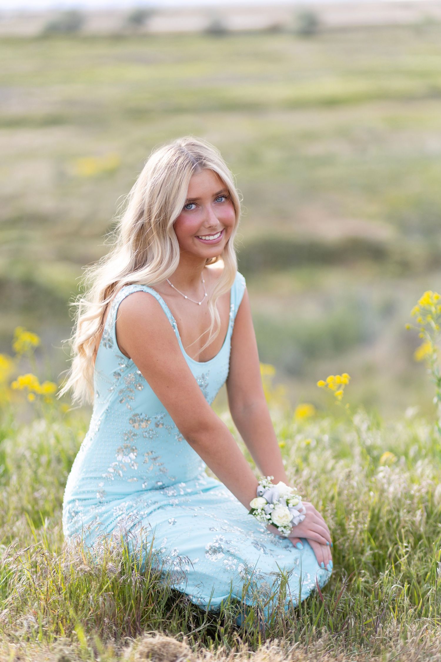 Smiling blonde woman in light blue floral dress sitting in a wildflower field holding small white flowers.