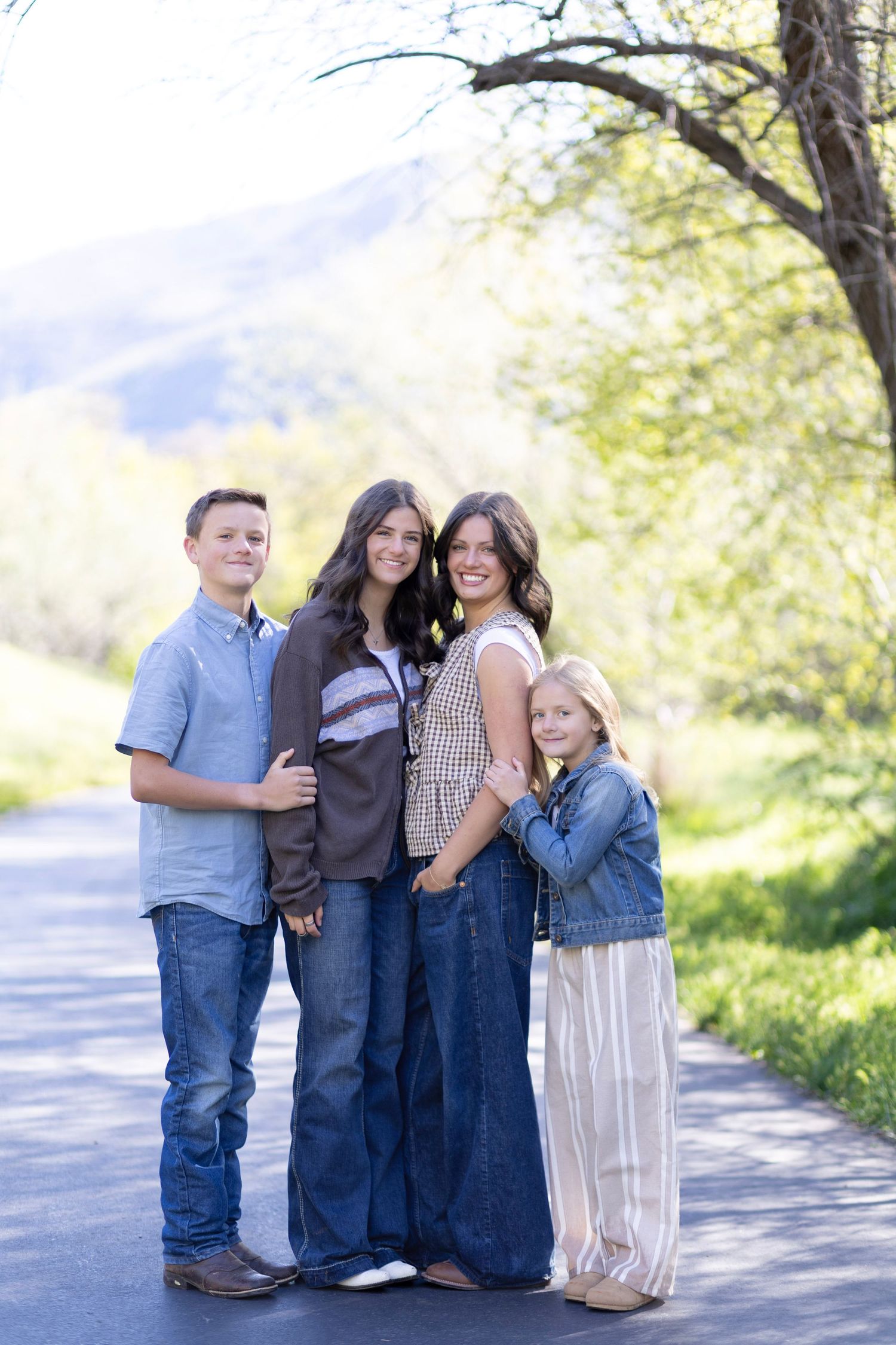 Family of four posing on a scenic road surrounded by lush green trees and mountains in the background.