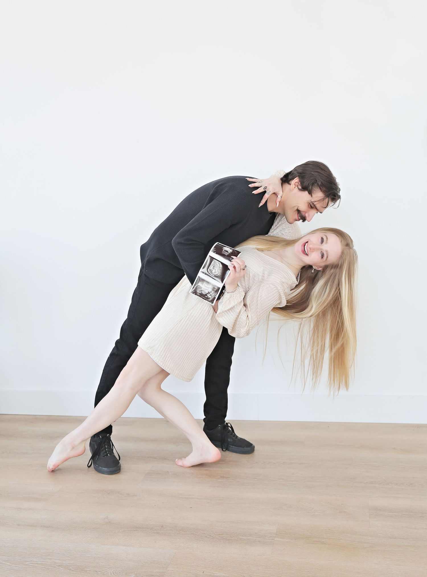 Playful couple strikes dramatic dance pose while holding ultrasound photos in bright indoor setting.