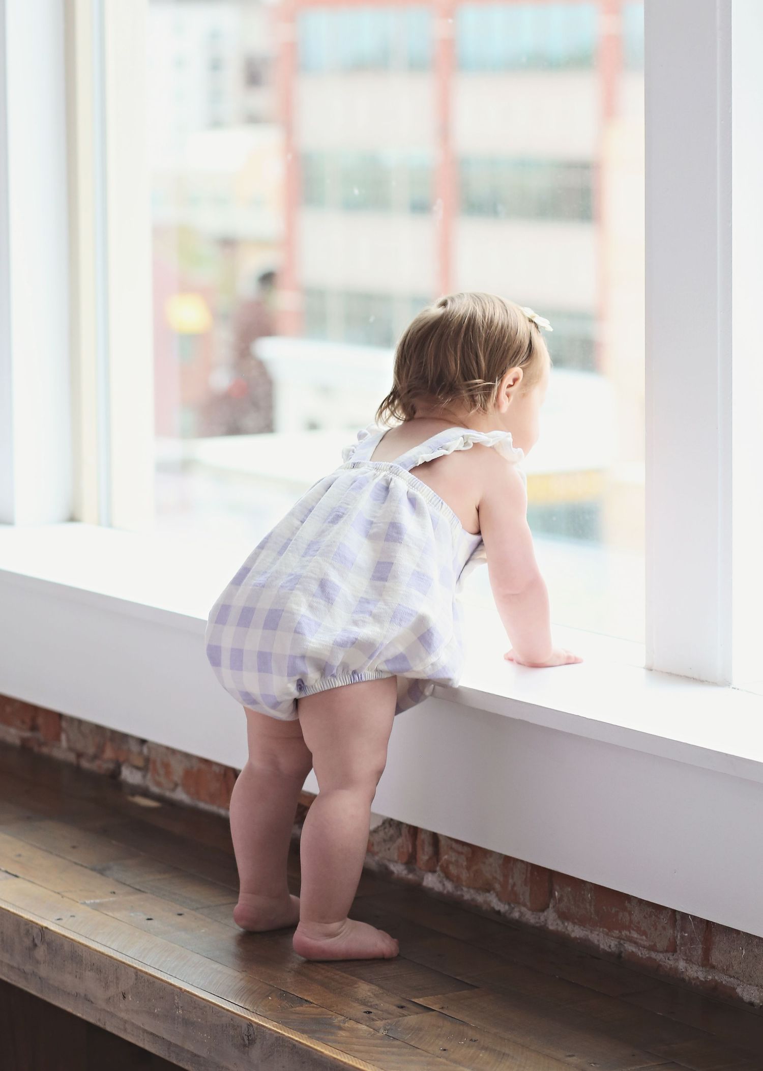 A toddler in a gingham romper stands on tiptoes looking out a bright window.